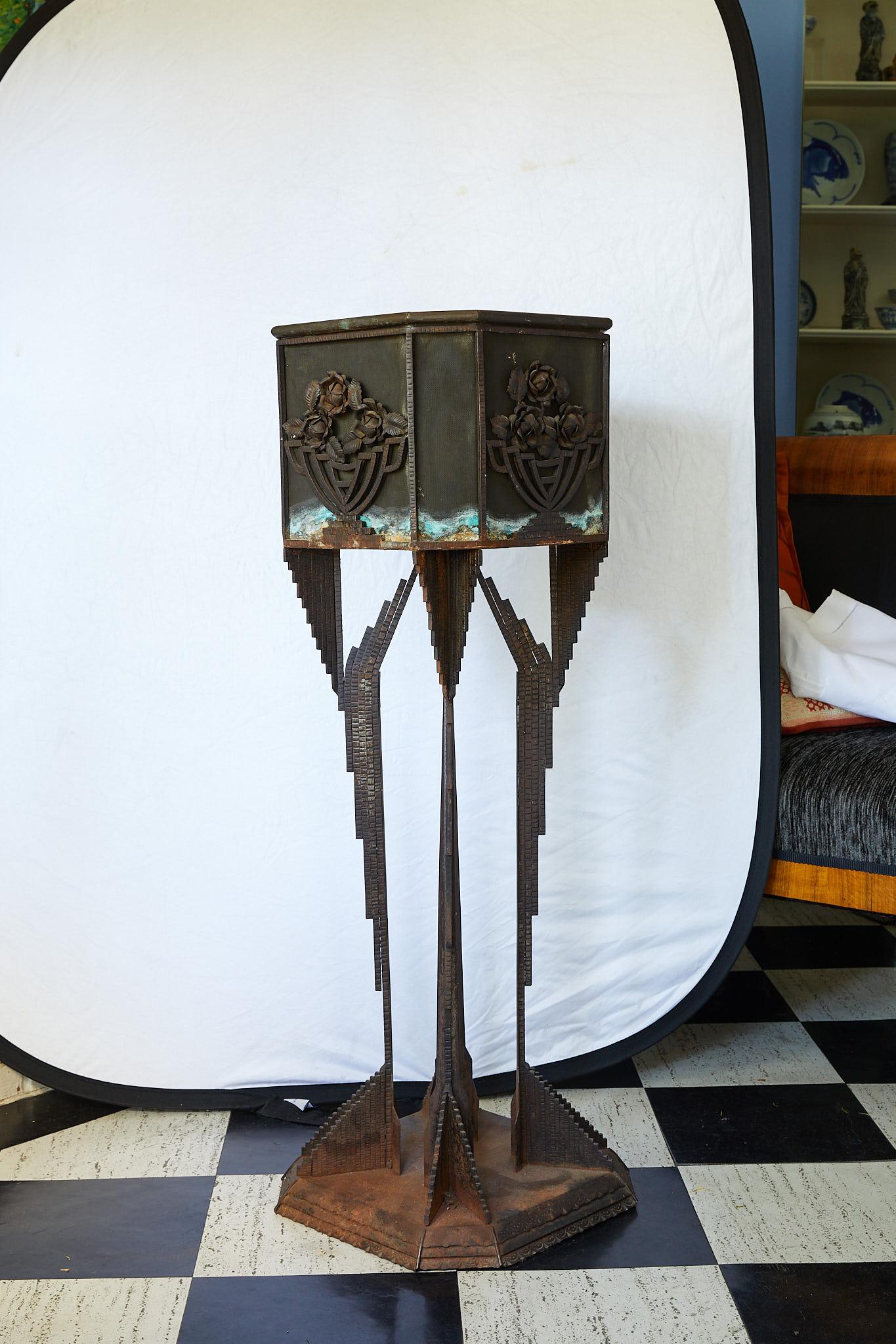 Art Deco sculptural iron pedestal supporting a square wire mesh planter with decorative appliqués of metal flowers in iron bowls. The planter holds a metal liner on its interior. The pedestal is made of four cast iron winged legs on a tiered