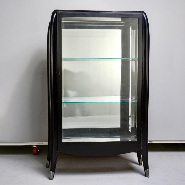 Italian Art Deco glazed display cabinet features an ebonised finish with curvy lines and silver toned metal sabots, circa 1930s. Cabinet has functional lock on door and two fixed internal glass shelves. Shelves measure: 29.75” wide x 11.75” deep.