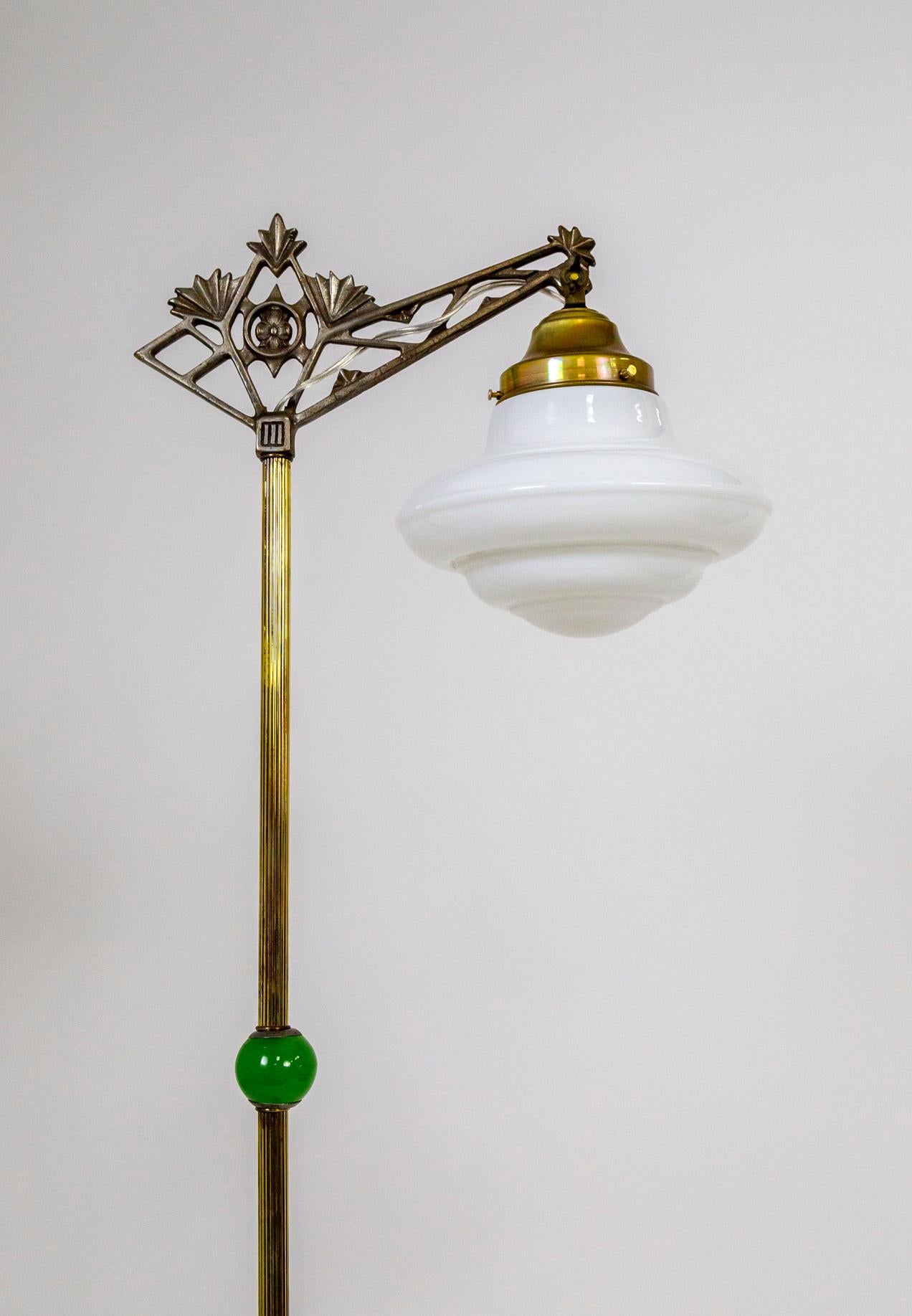 An early 20th century floor lamp with a reeded, brass stem and ornate, geometric crown/bridge that holds a rounded, tiered, milk glass shade. With jade green glass accented in the middle of the stem and stacked on the beautifully patinated, cast