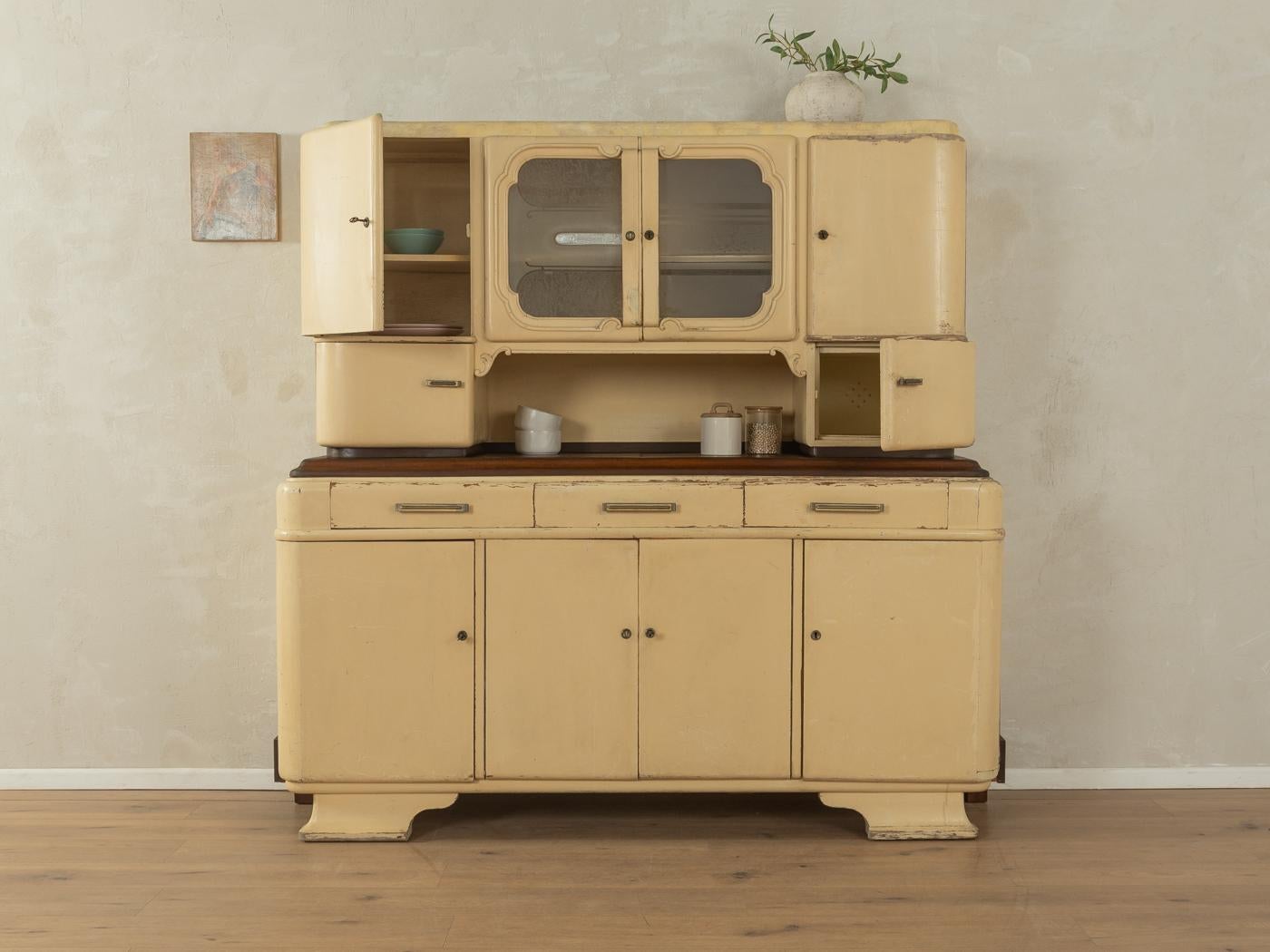 Wonderful two-part Art Deco kitchen cabinet from the 1920s. High-quality corpus made of blockboard with eight doors, two glass doors, six shelves, three drawers and period-typical feet. Designed and manufactured in Germany.

We thoroughly