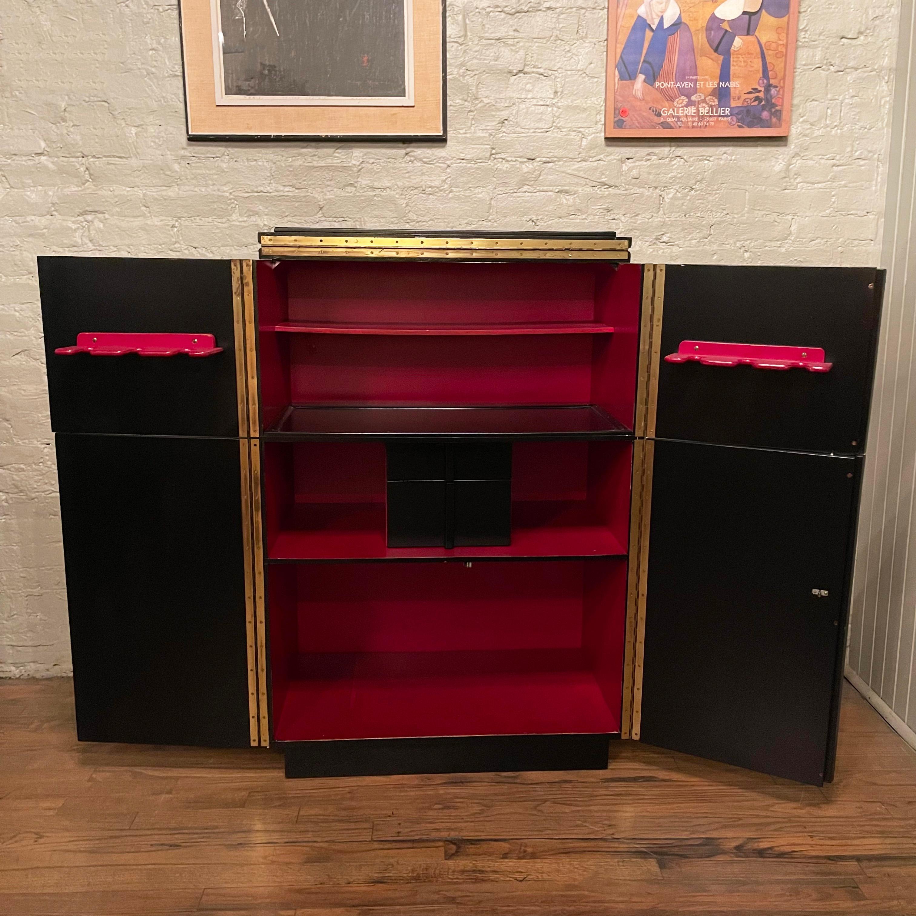 Streamlined, art deco / midcentury, walnut cabinet with a black lacquered exterior opens to a 60 inch wide full bar with contrasting lipstick red interior finish. The interior features open shelves for bottles and glasses, two small drawers, slots