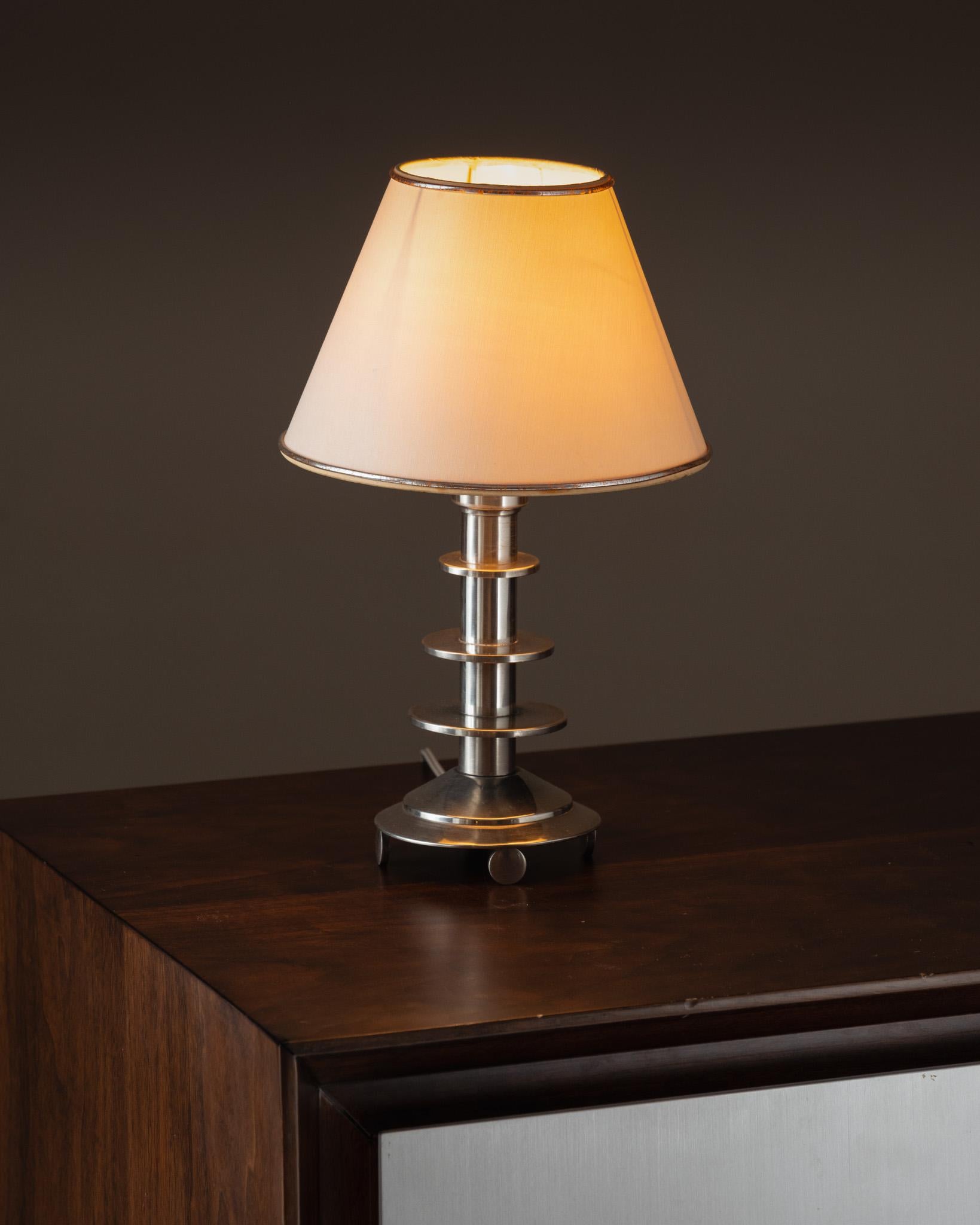Petite lampe Art déco, années 1930. Son pied en étain poli, rythmé par une succession d’anneaux cylindriques, est typique de l’époque. Très bel état vintage, fonctionnelle, équipée d’une prise européenne. Vendue avec son abat-jour d’origine.