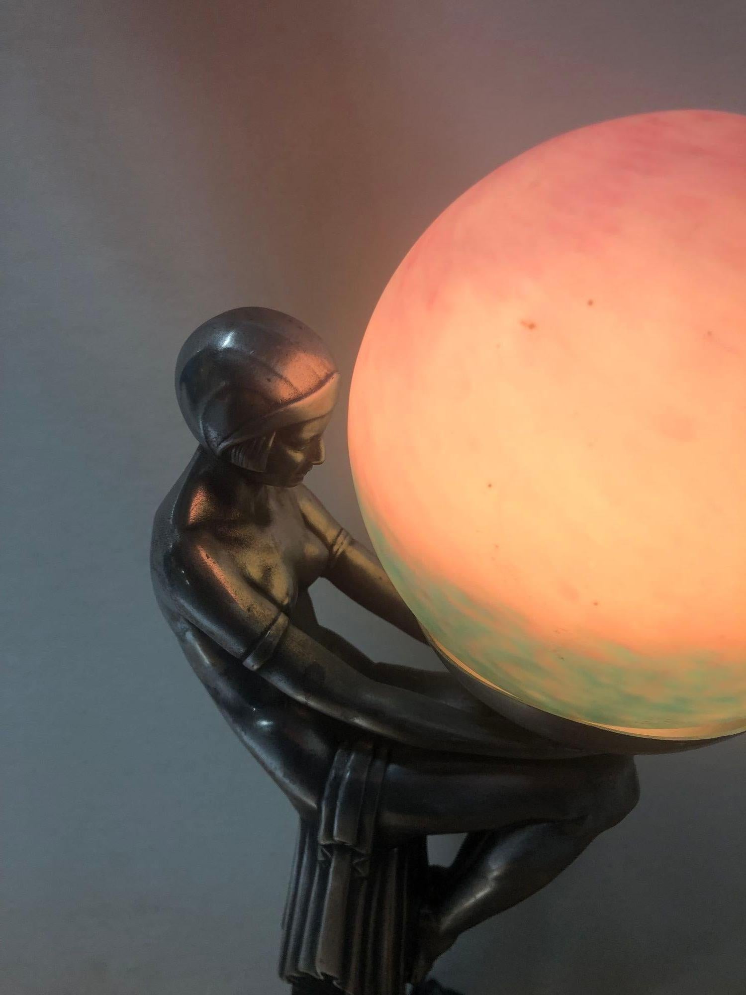 A tribute to the Art Deco era, the 1920s lady gracefully holds a large green and pink globe bulb, reminiscent of Max Le Varrier's iconic style. Adorning a stunning grey marble pedestal base, this embodiment of elegance and sophistication captures
