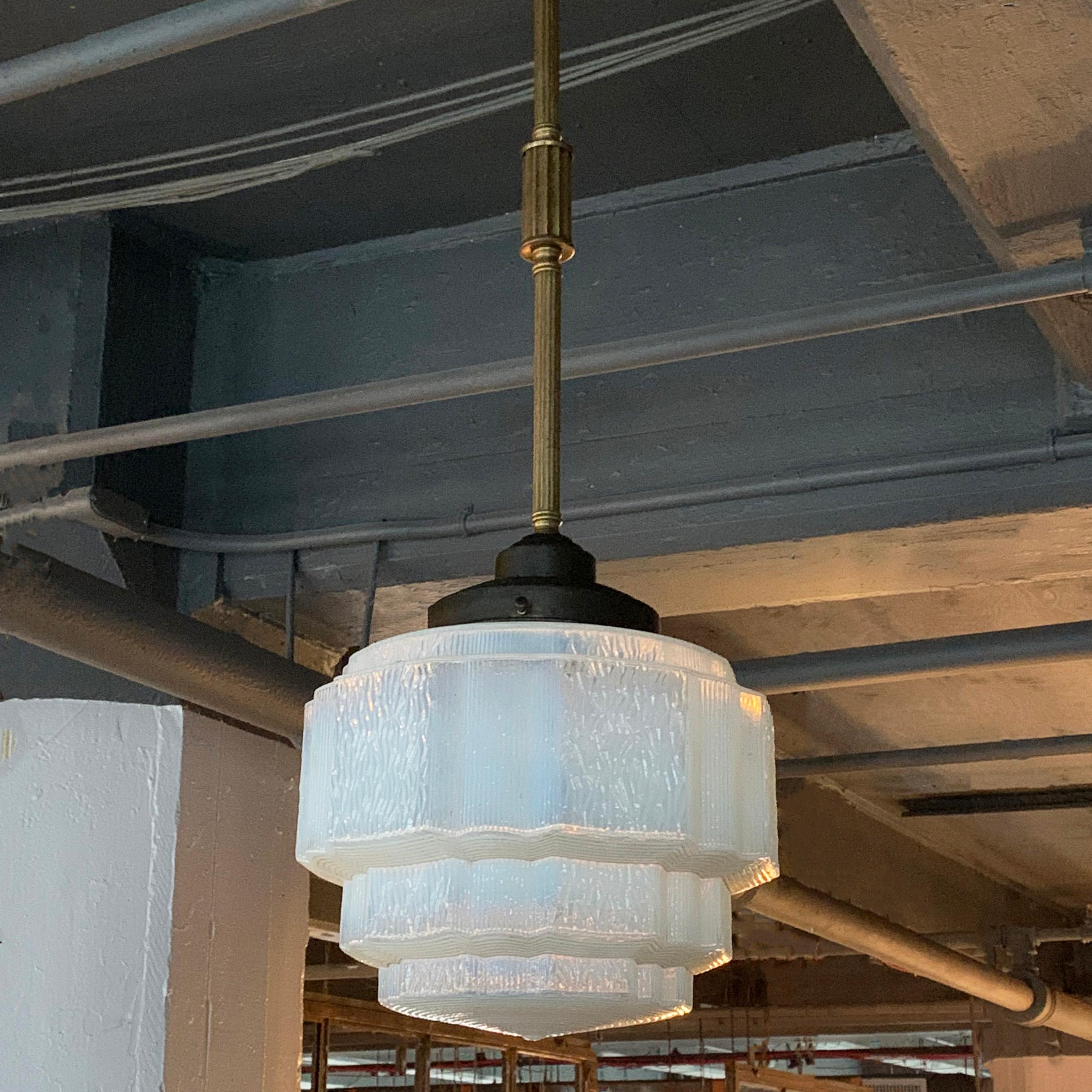Art Deco pendant light features a layer cake, ribbed pattern opaline glass shade on a decorative, steel and brass pole with canopy. The pendant is newly wired to accept up to a 200 watt medium socket bulb. The shade measures 12 diameter x 9 height.