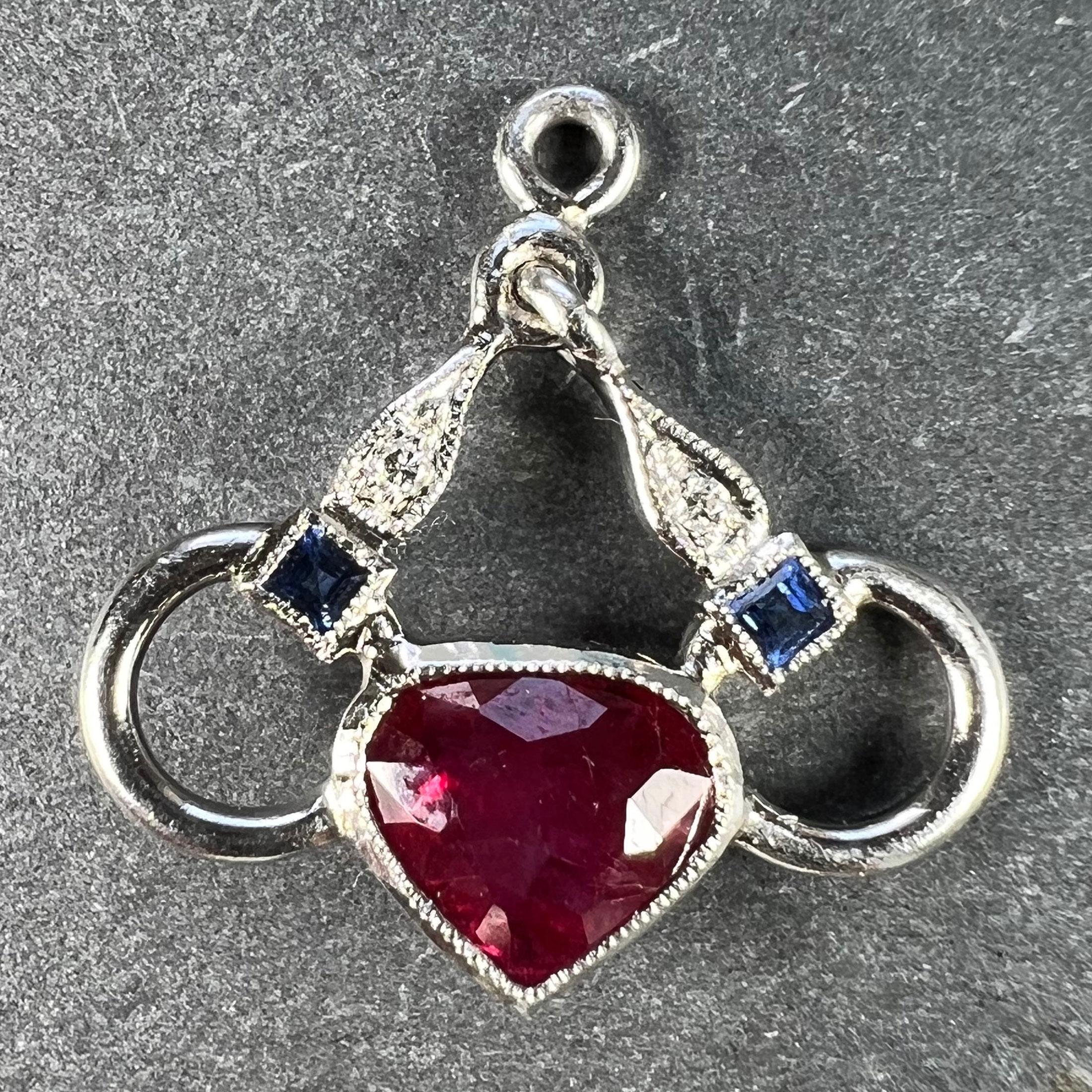 An Art Deco platinum charm pendant designed as a snaffle bit for horse riding with a ruby love heart and a sapphire and diamond set bit. The charm is set with a fracture-filled ruby heart, two round brilliant cut diamonds and two carre-cut