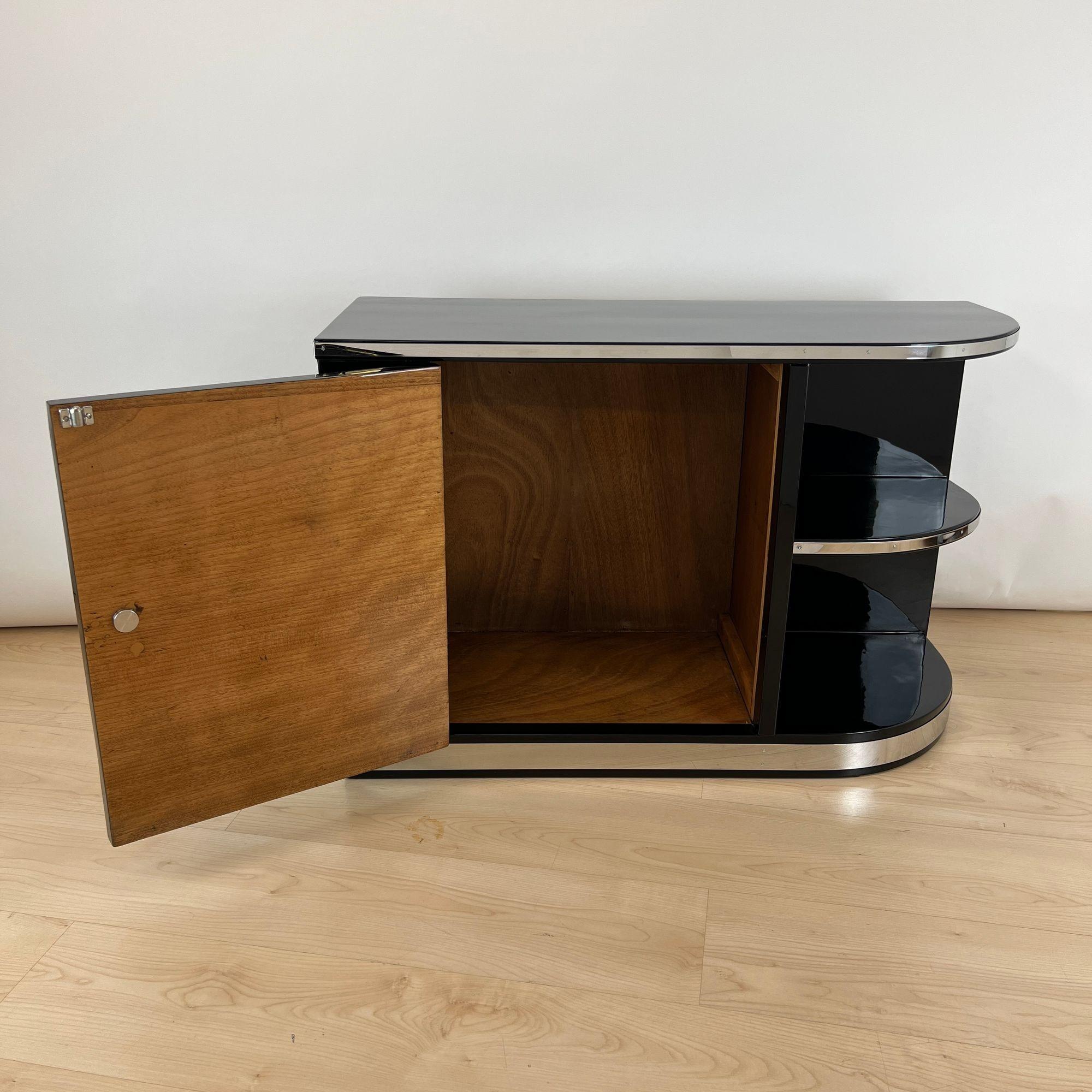 Small asymmetrical Art Deco Lowboard from France around 1930.
Black, high-gloss polished piano lacquer on wood. Door with chrome-plated knob, turned rosette and snapping ball lock. Inside in walnut, on the inside of the door in bright lacquered