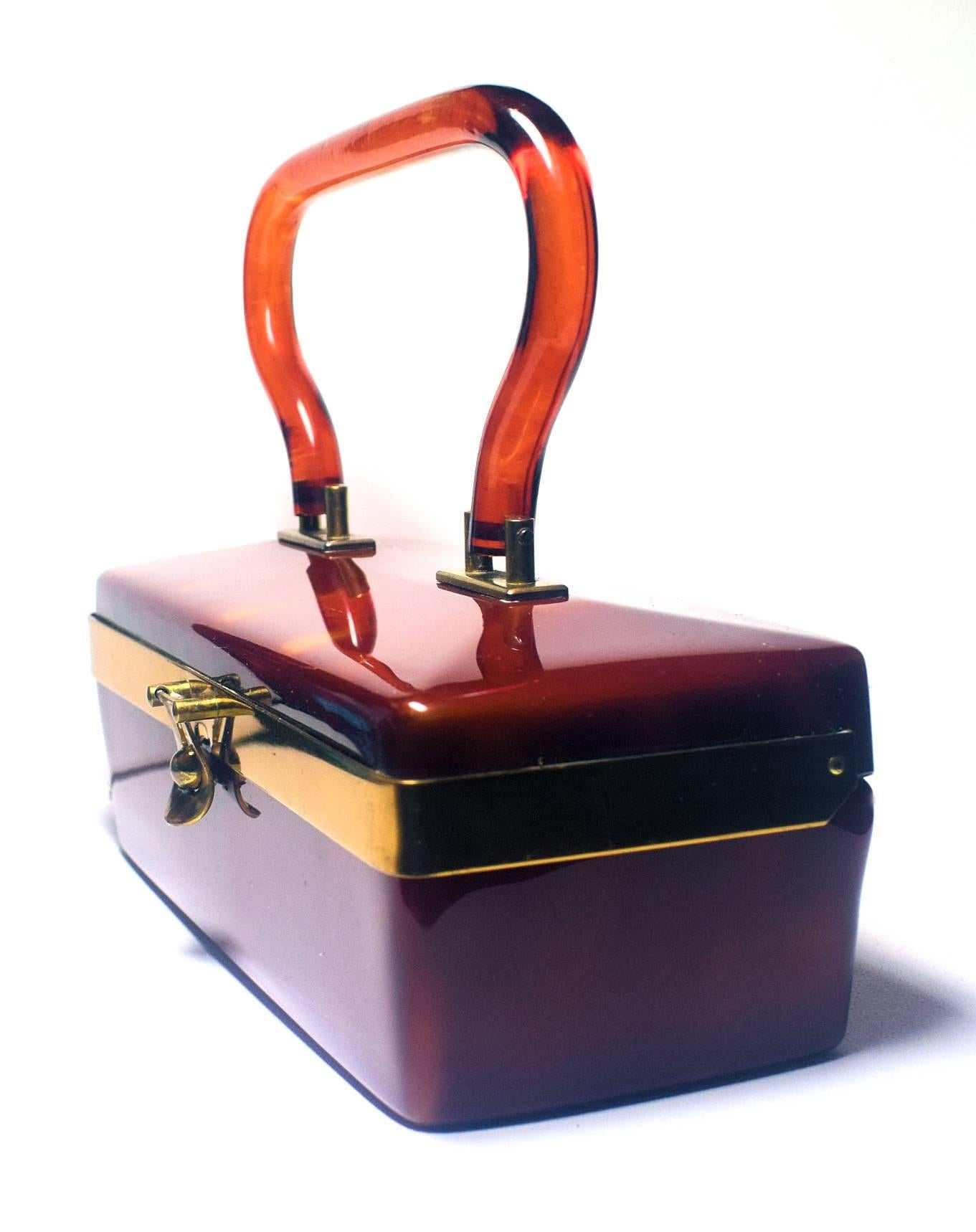A beautiful, glossy caramel Lucite box bag. Unknown manufacturer, there are the remains of a brand on the mirror, but it is illegible. The box is completely made from a dark swirled caramel Lucite and has a brass frame.
Absolutely stunning and very