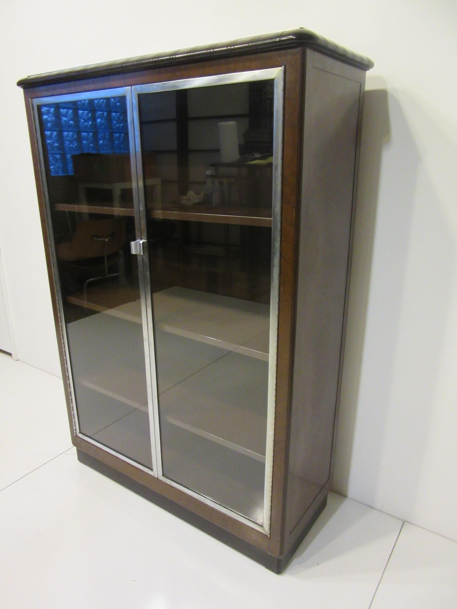 A wonderful Art Deco streamline machine age bookcase having a faux wood painted metal cabinet, walnut top, Bakelite type top trim edge with detailed groove design. The front has two glass doors surrounded by aluminum trim and matching handles