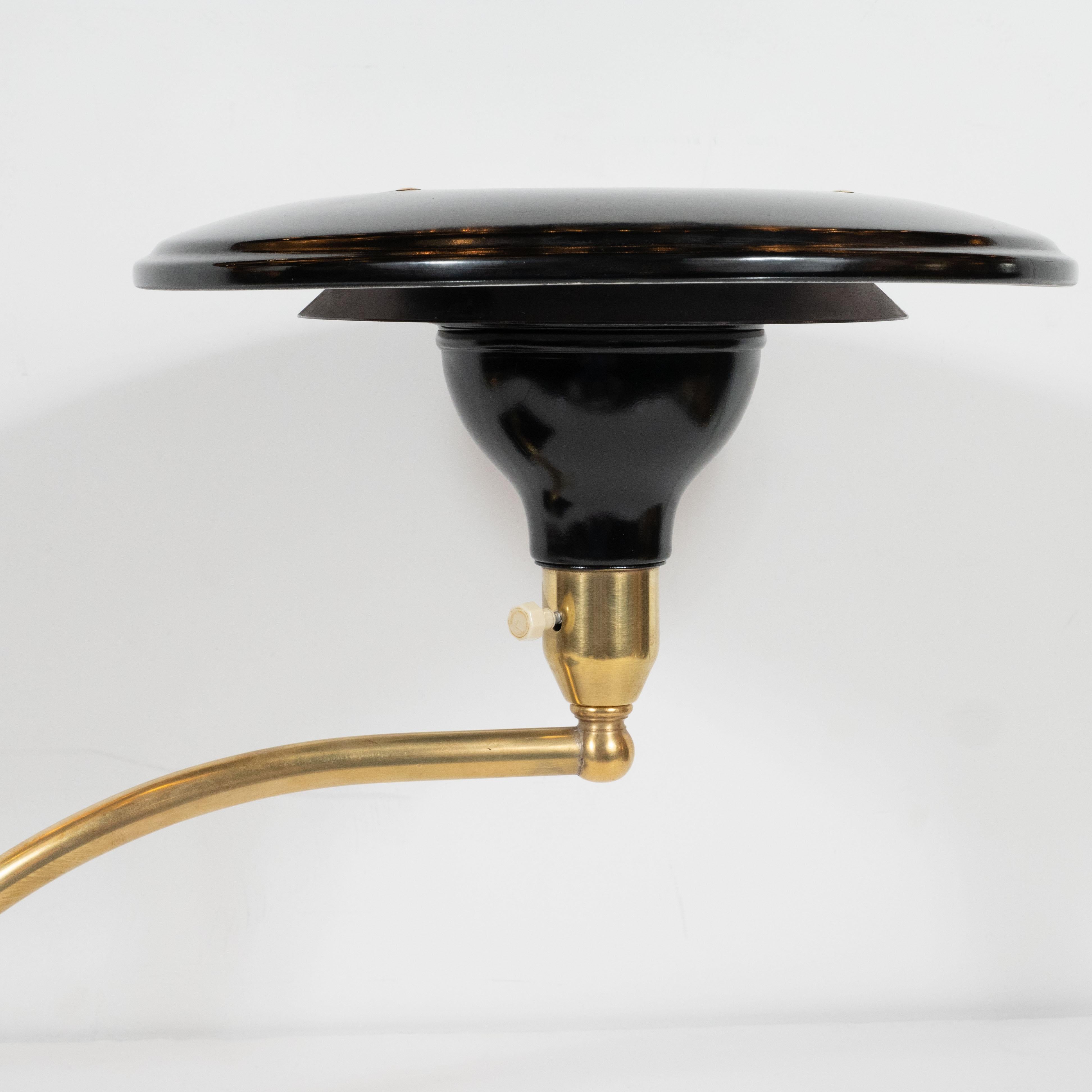 This elegant Art Deco Machine Age table lamp was realized in the United States, circa 1935. It features a rectangular black enamel base plate topped with a spherical brass embellishment. A curved arm extends outwards culminating in a conical brass