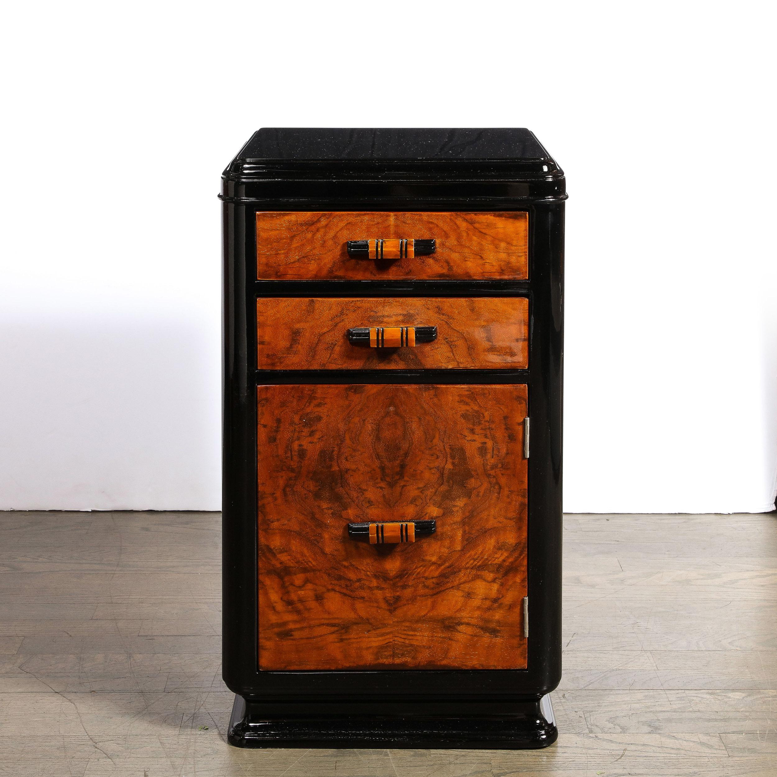 This elegant Machine Age Art Deco nightstand was realized in United States circa 1935. It features a lustrous black lacquer base and body with two top drawers and a lower compartment in stunning bookmatched burled walnut (showcasing the rich natural