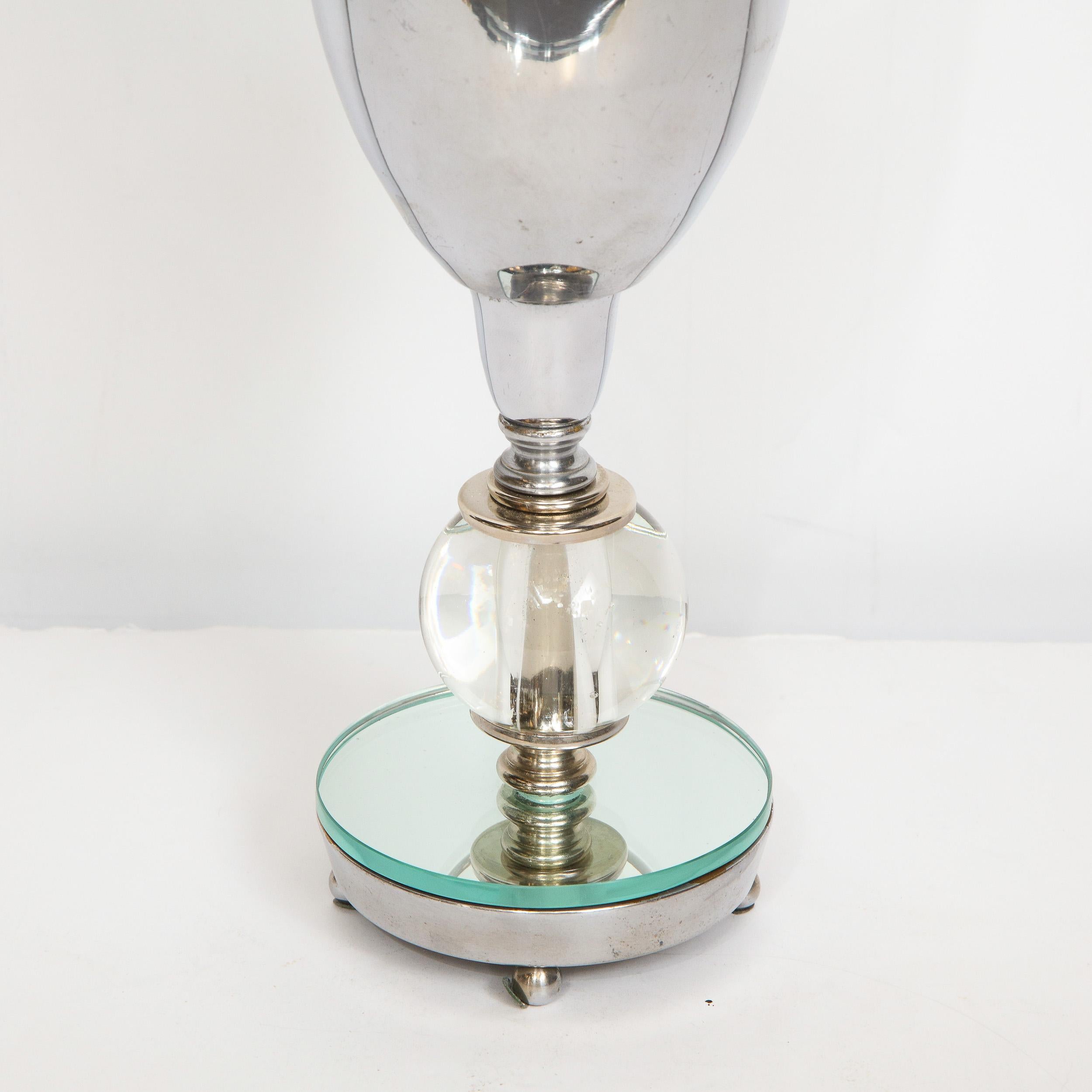This glamorous Art Deco Machine Age uplight was realized in the United States, circa 1935. It features a chrome and mirrored base sitting on four ball feet. The body offers a translucent orbital embellishment in the center topped with skyscraper