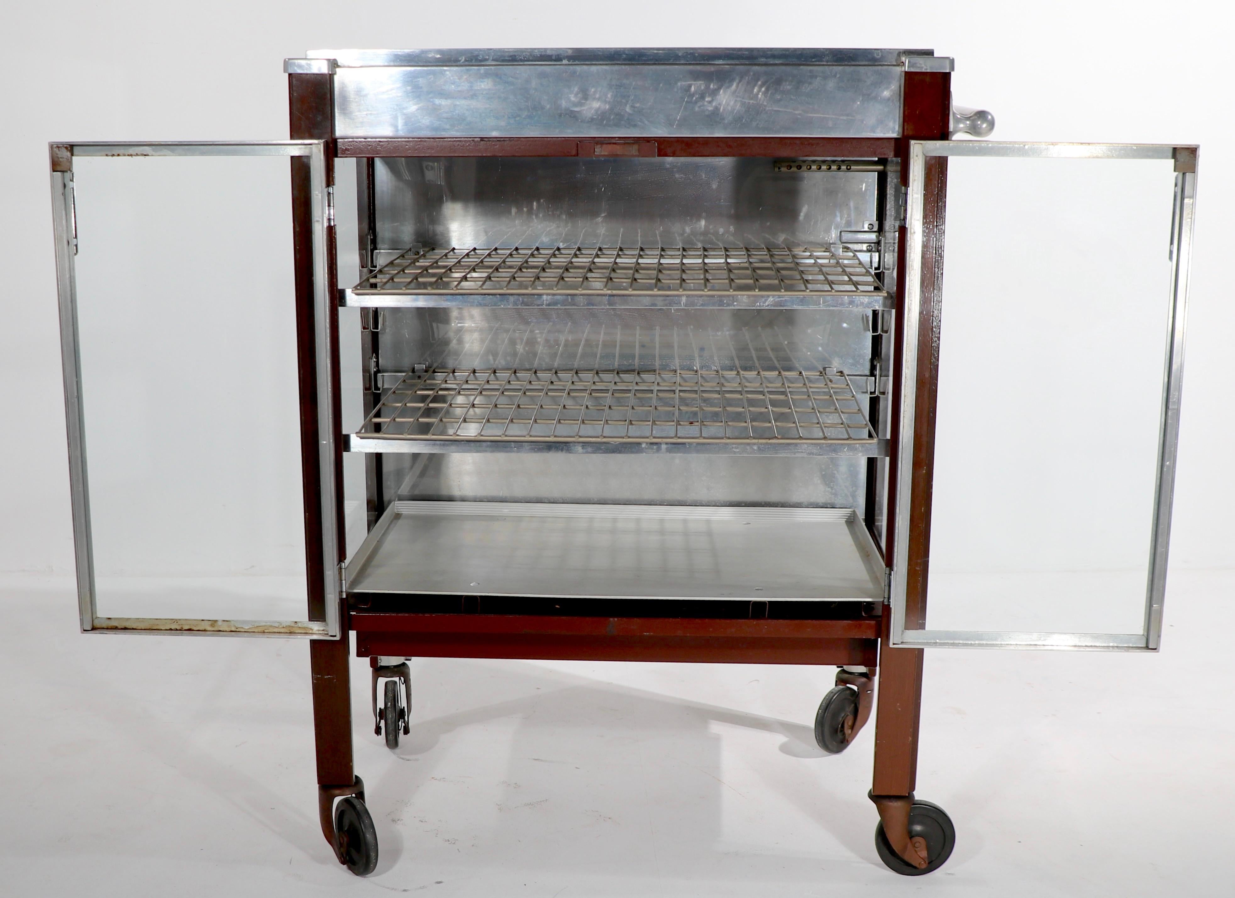 Fabulous Art Deco Machine Age serving cart, having a glass enclosed center, with a heated surface, and a heated top surface. Center case has two swing out doors, and two adjustable shelves. The case is solid wood, and the frame is steel and cast