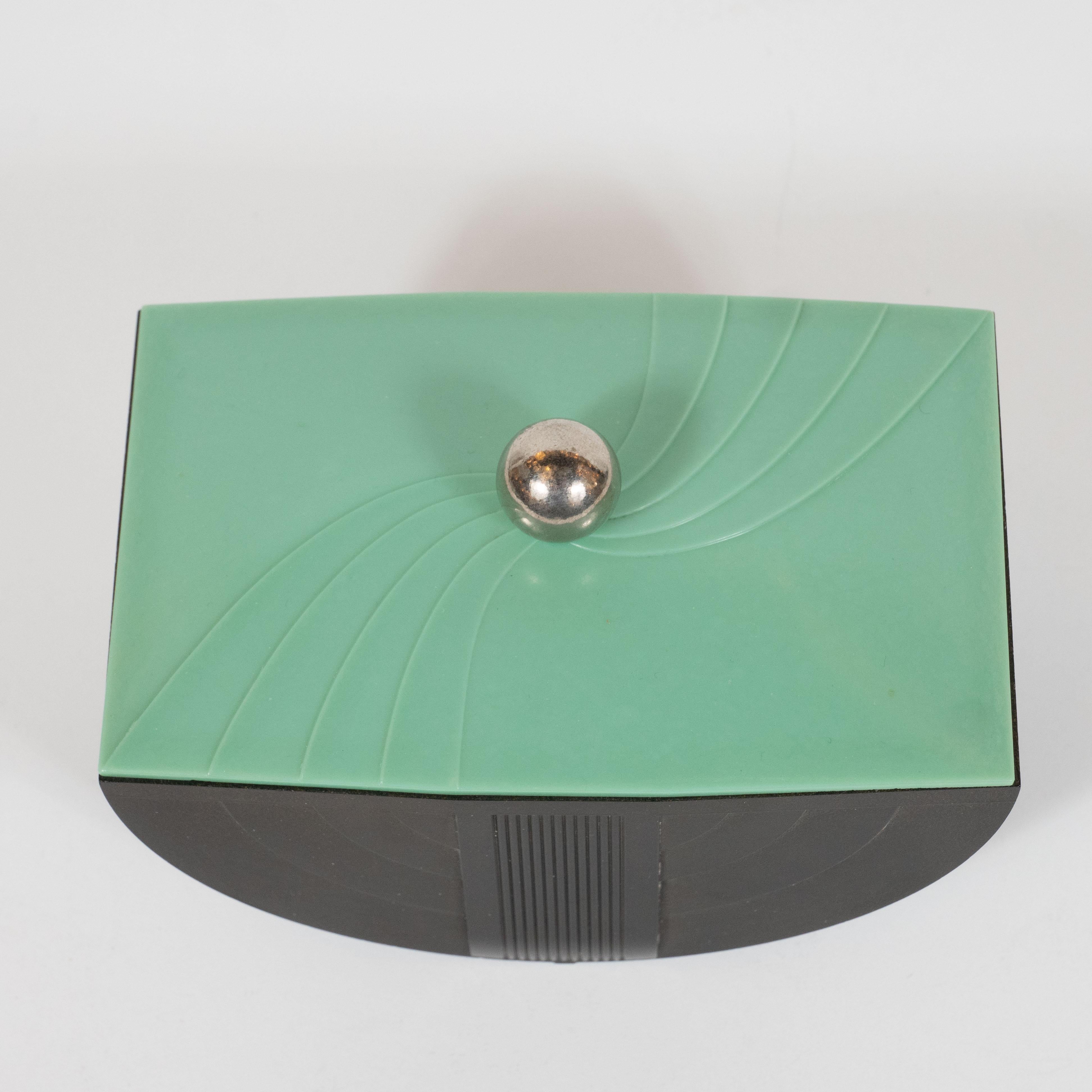 This elegant machine age Art Deco Bakelite box was realized in the United States, circa 1935. Sitting on spherical chrome feet, this stunning box features a demilune body in black Bakelite with streamlined designs tracing the demilune form of the