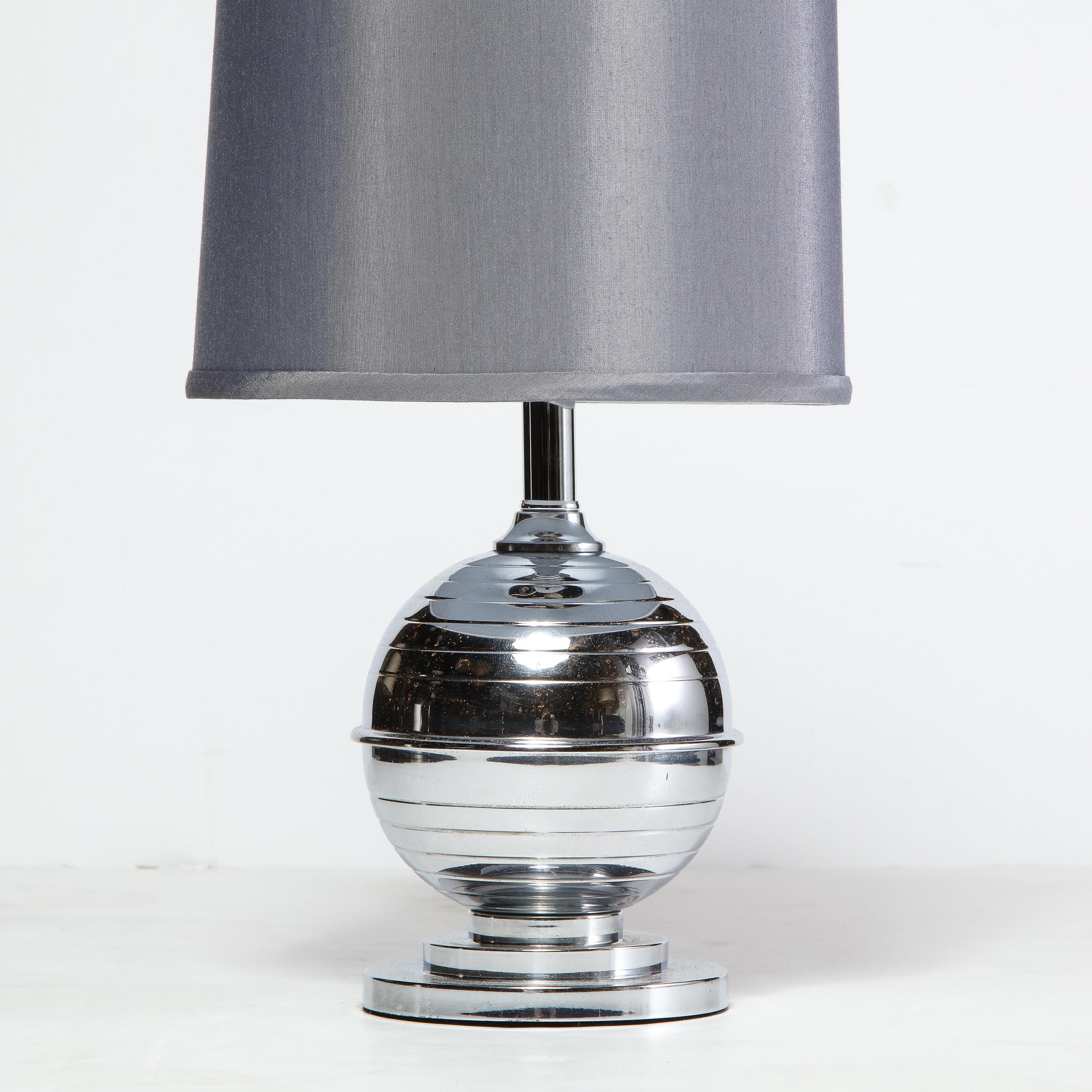 This stunning Art Deco Machine Age table lamp was realized in the United States, circa 1935. It features a two tiered skyscraper style base that ascends into a spherical banded body in polished chrome with a tapered neck. With its clean modernist