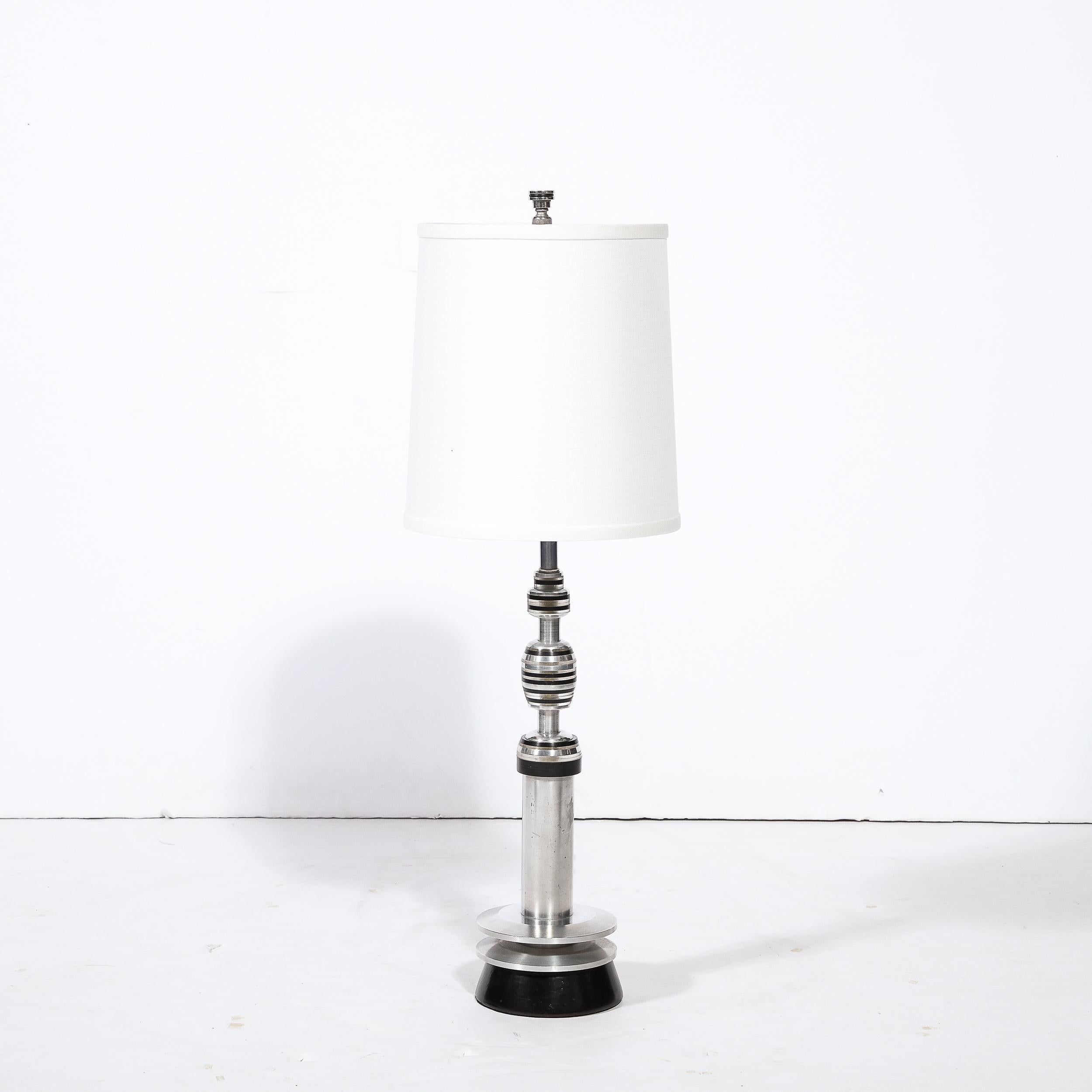 This refined Art Deco Machine Age table lamp was realized in the United States circa 1935. It features a skyscraper style conical black bakelite base with two ringed embellishments floating above that are pierced through by the cylindrical aluminum