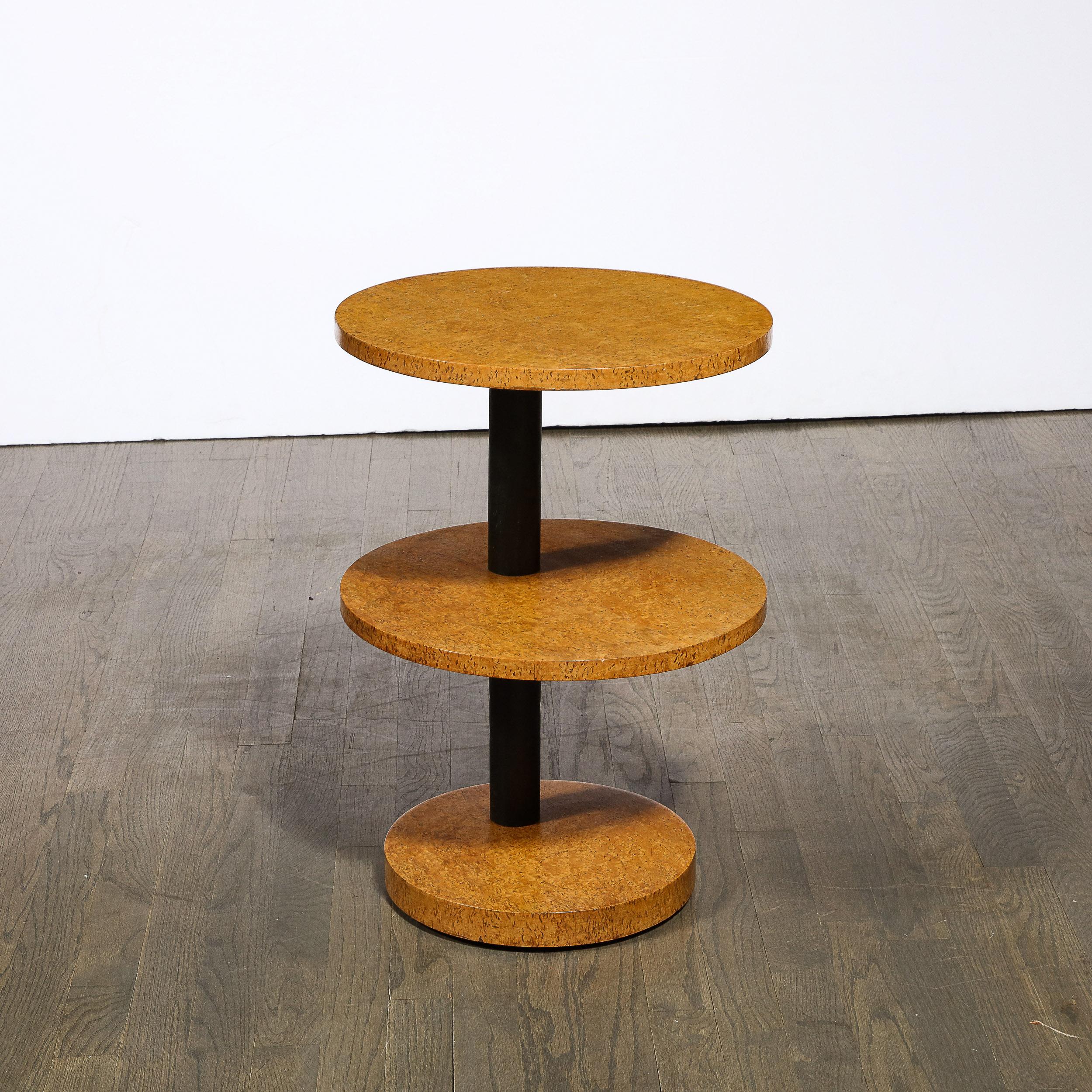 This stunning Art Deco Machine age three tier side/ end table was realized in the United States circa 1935. It features three circular volumetric tops in a beautiful burled elm (presenting a stunning natural blonde wood grain) pierced through by a