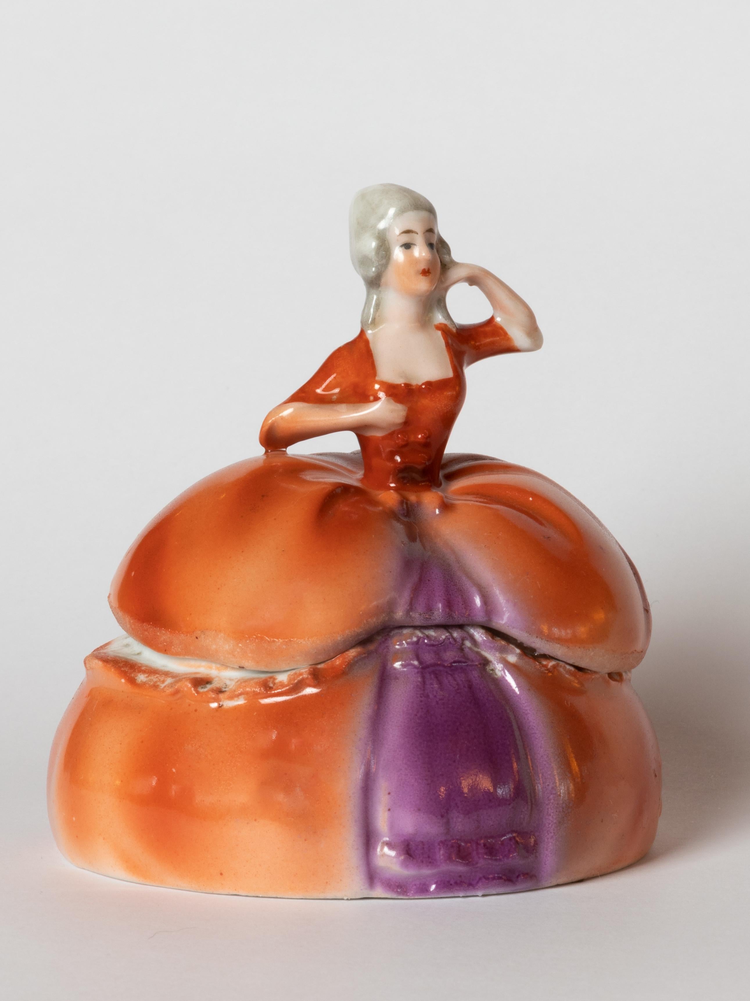 This Art Deco porcelain powder box, known as “Madame Pompadour,” was produced in 1929 by Schumann Arzberg in Bavaria, Germany, for Golden Crown E&R. 
Designed in the form of a fashionable 18th-century lady with powdered hair and a voluminous orange