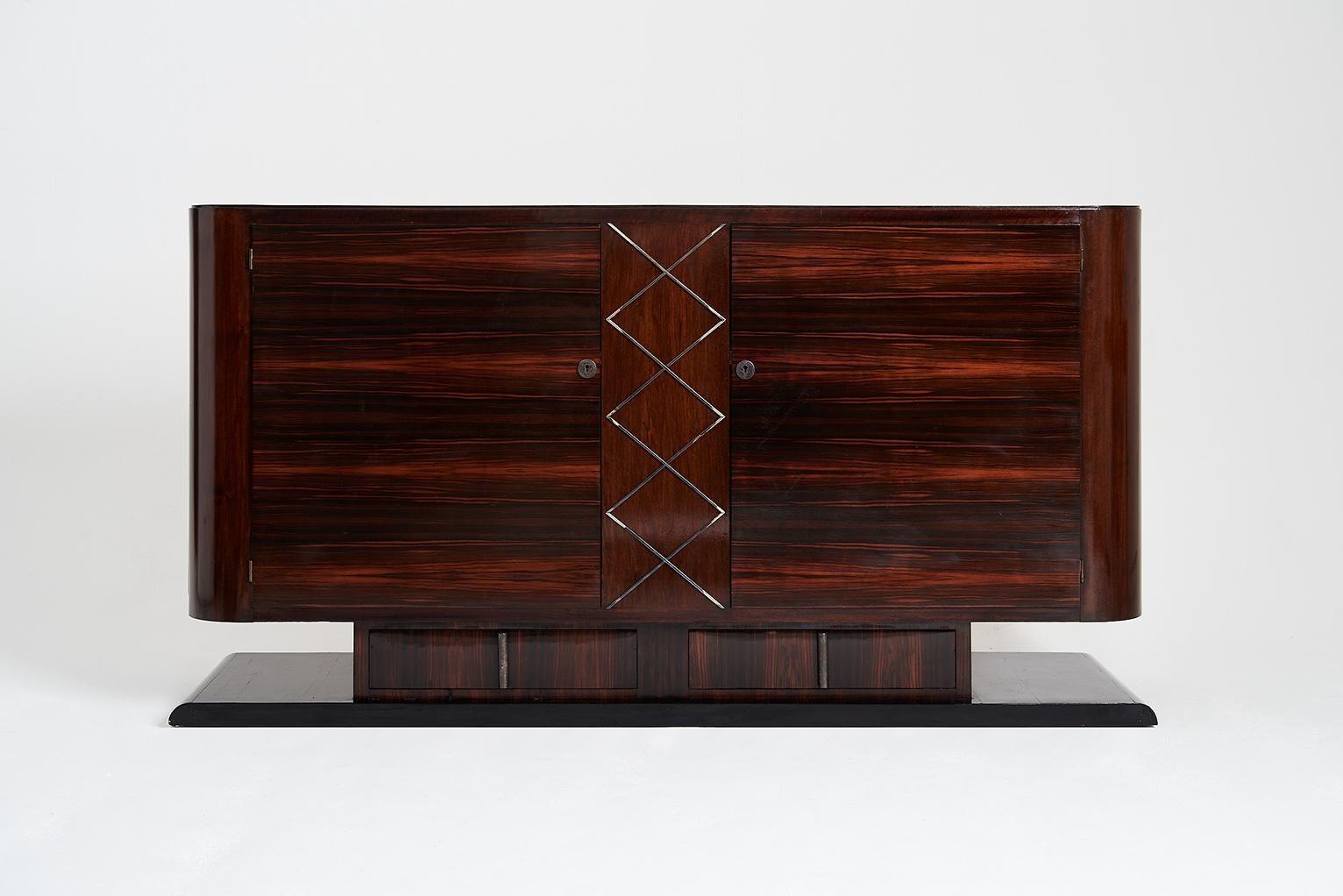 An Art Deco makassar ebony sideboard, internally oak lined, the ebonized plinth with two makassar ebony drawers, supporting a two-door body with rounded corners, centered by nickel geometric mounts.
France, circa 1930.