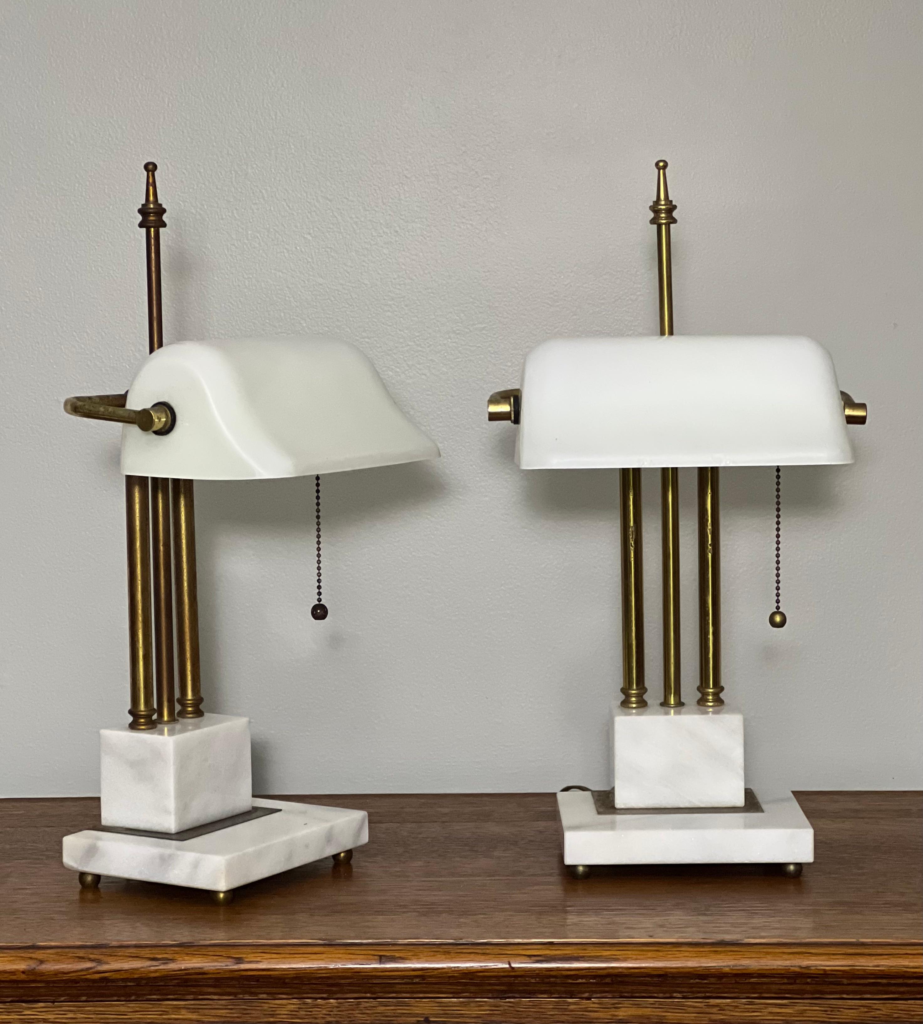 Unique pair of Art Deco brass and marble bankers lamps with hand-blown white milk glass shades, c. mid 20th century. They feature a tilting shade with pull chain, a triple brass bar stem and tall finial set upon a white marble base with brass ball