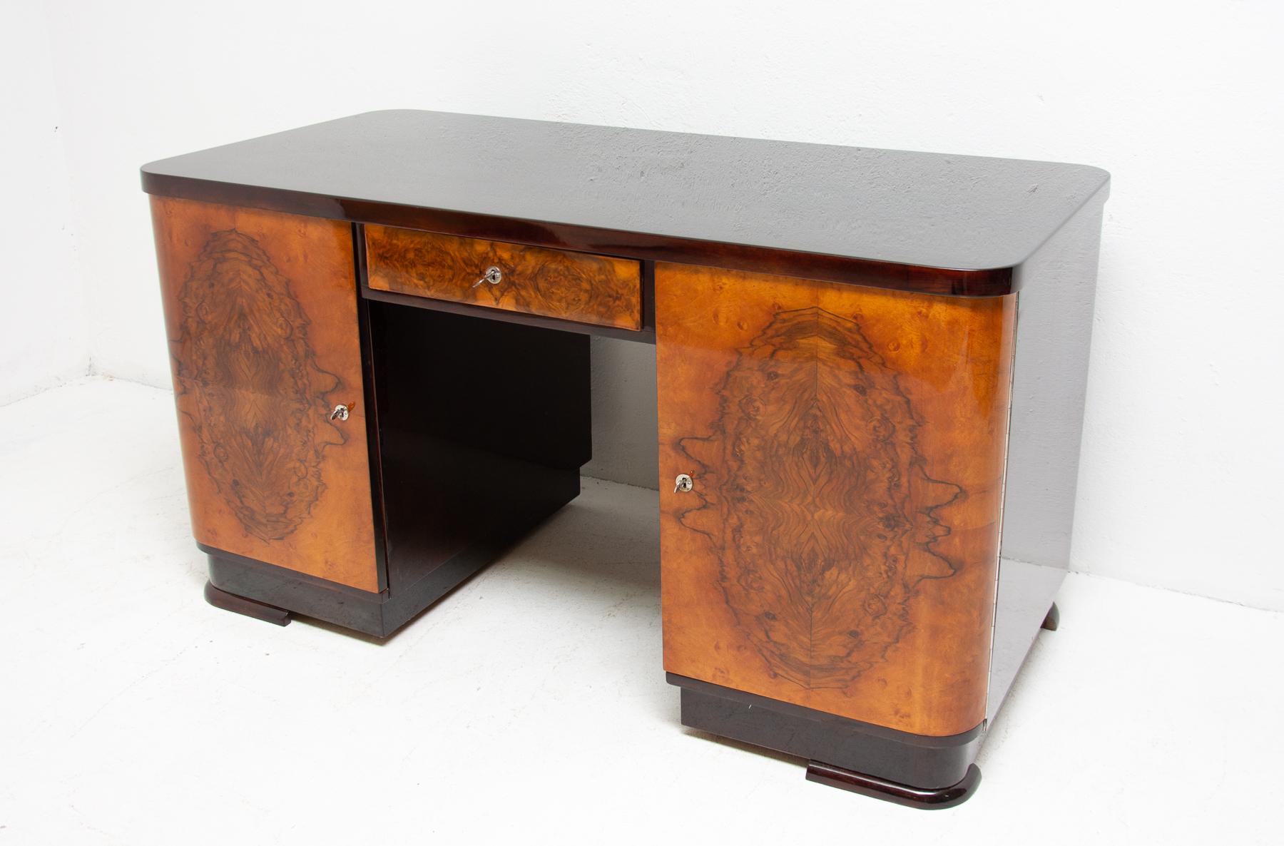 This massive Art Deco writing desk was made in Bohemia in the 1930s. It´s made of solid wood and features a walnut veneer inlaid. The top is made of oakwood. It includes one drawer and four extended shelves. The desk is in excellent condition due to