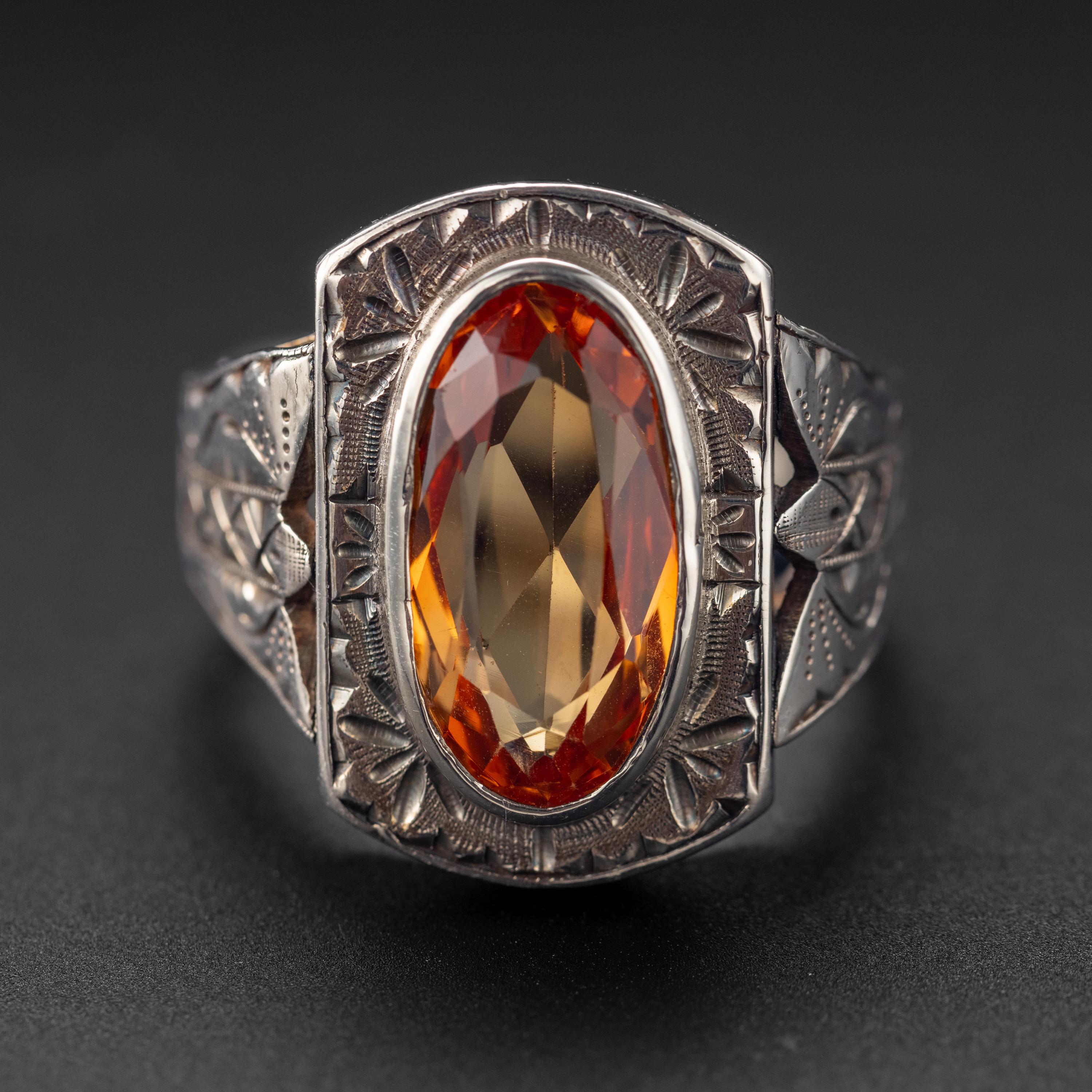 This bold and handsome hand-made ring from the 1930s features an 18.50mm x 10.13mm oval-cut orange sapphire weighing 7.62 carats set into a deeply engraved heavy silver mounting. The sapphire was created by the flame fusion process, invented in the