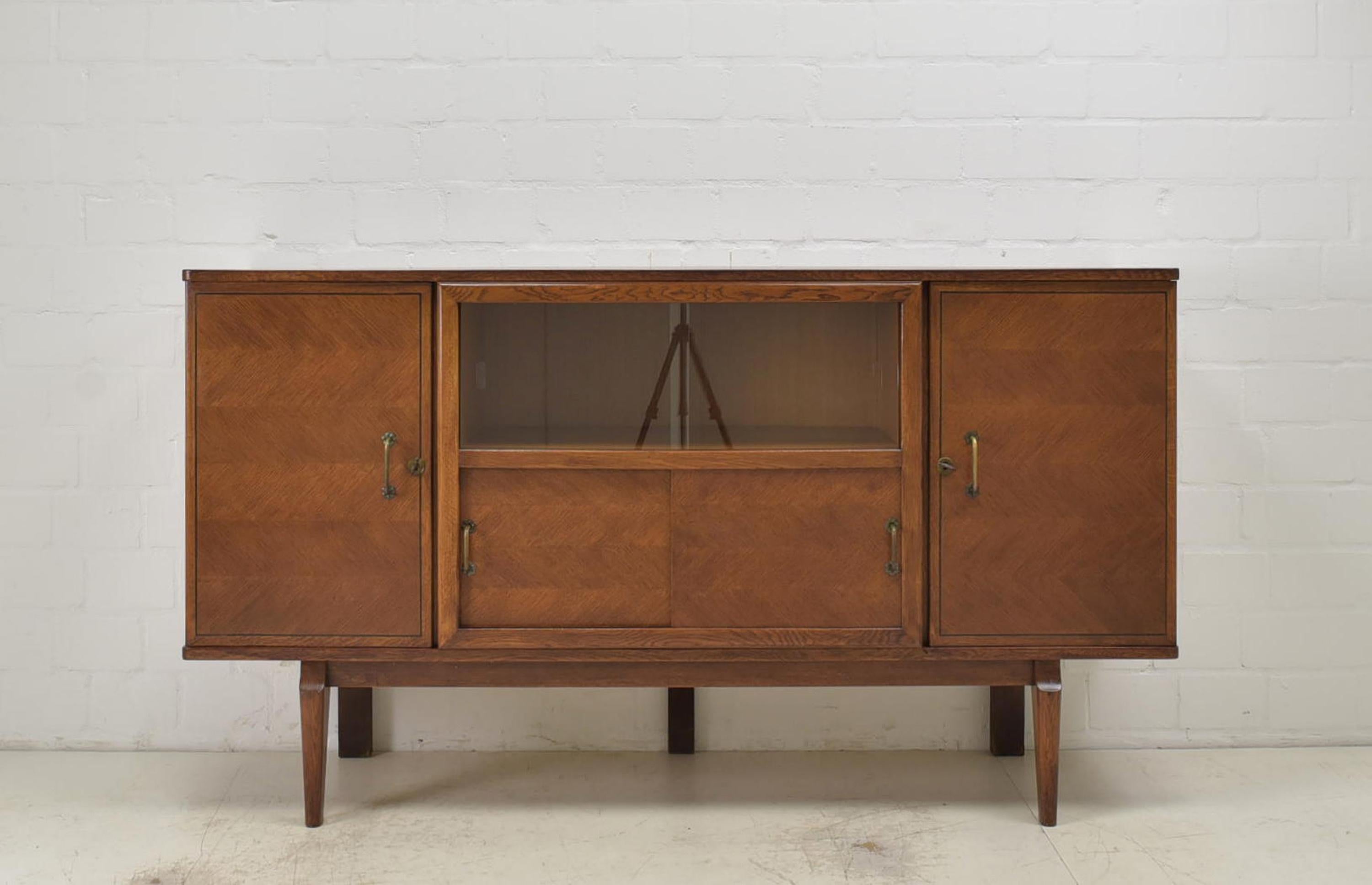 Showcase sideboard restored Art Deco midcentury around 1950 vintage oak

Features:
Two-door model with two sliding door compartments
One shelf each on the left and right, one with a drawer underneath
Very high quality processing
Shelves with