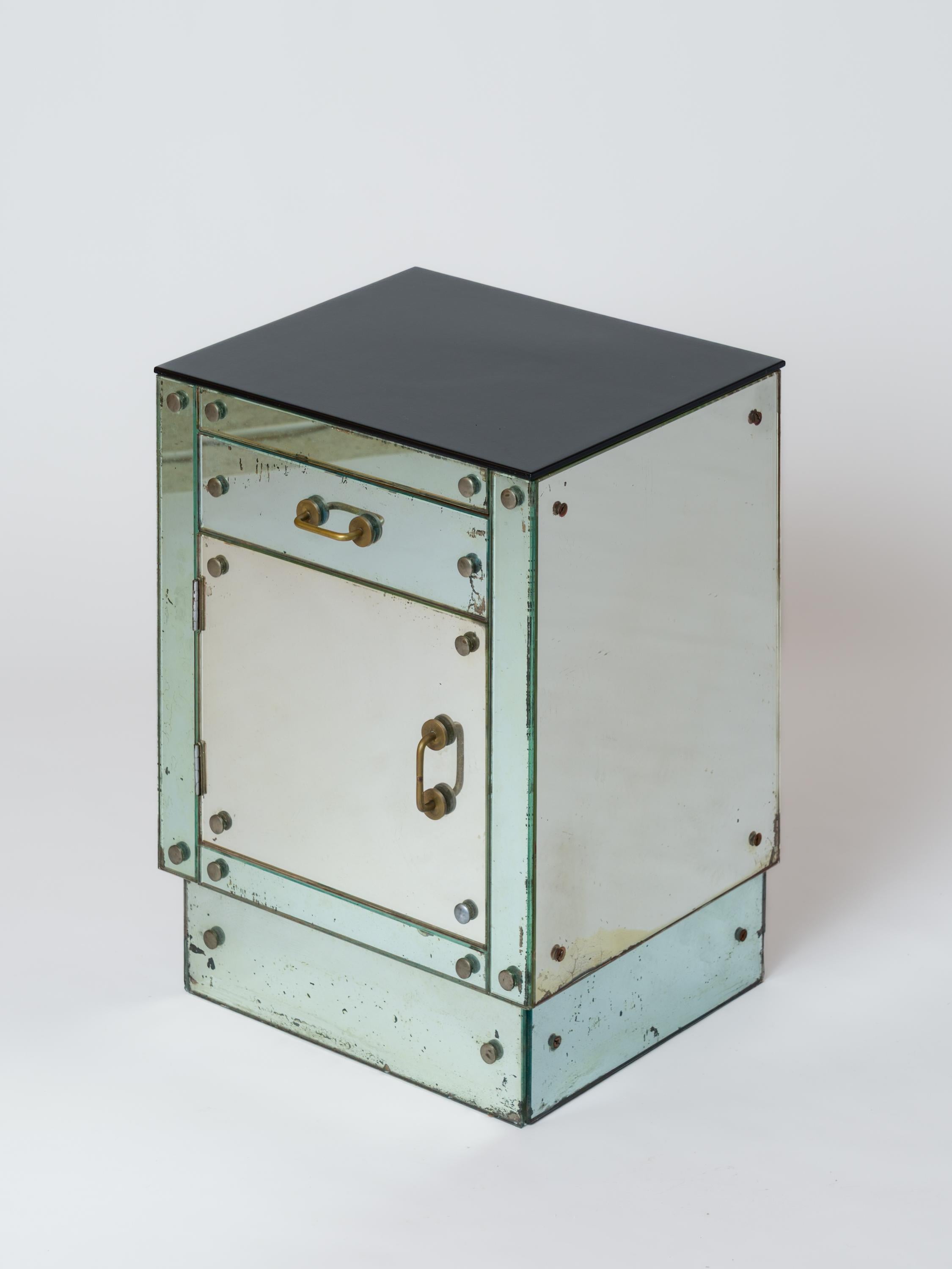 Un piccolo comodino rivestito di pannelli a specchio, con un cassetto e un vano inferiore con un'anta. I pannelli di vetro sono fissati con viti in acciaio nichelato. Le due maniglie sono in ottone patinato.
La superficie superiore è stata rifinita