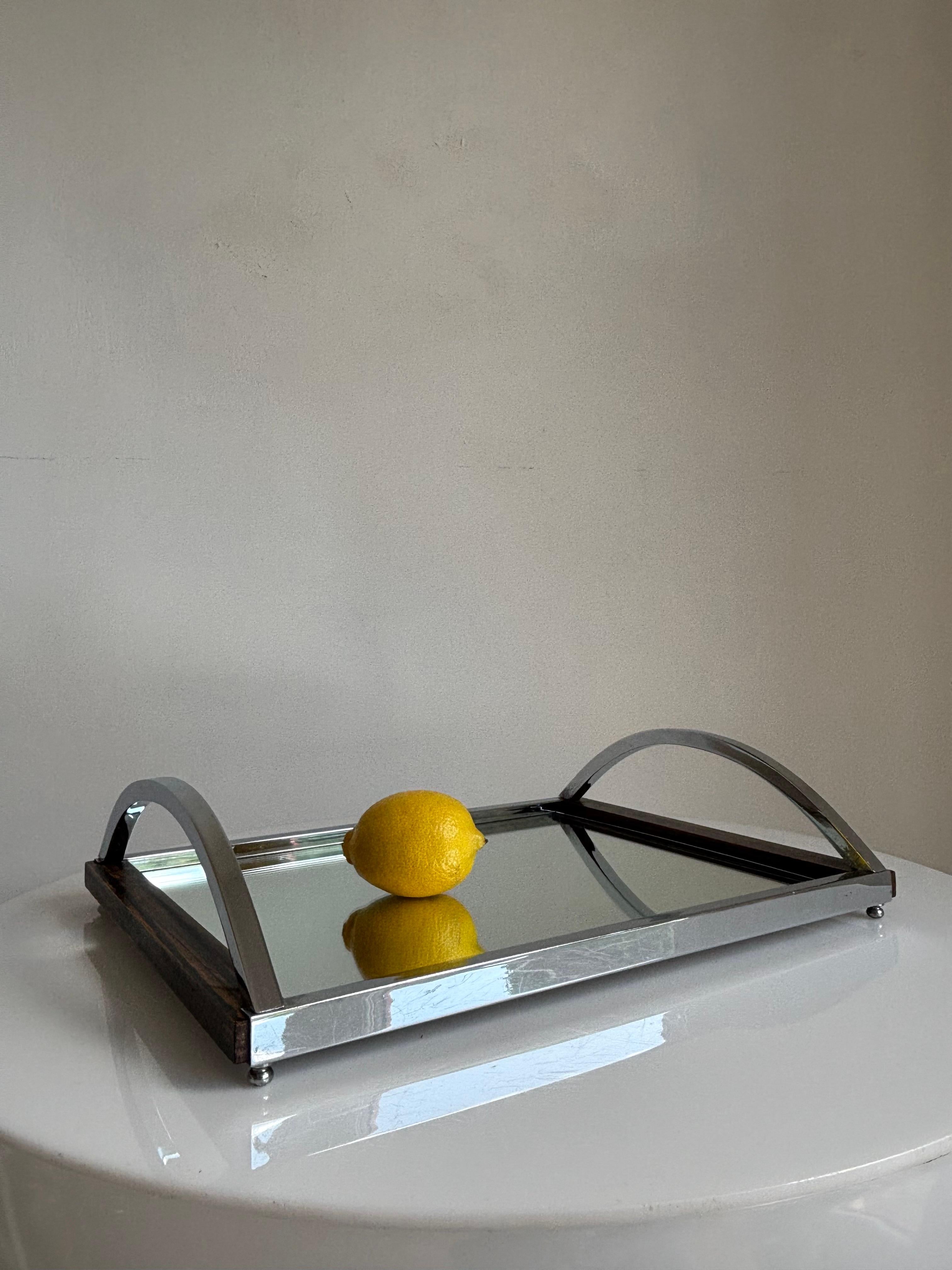 Gorgeous art deco serving tray composed of dark wood, shiny chrome metal and a mirror top. Four spherical feet under each corner and two large arched handles. 
A stunning decorative piece with a clean classic look that is easily used in any setting.