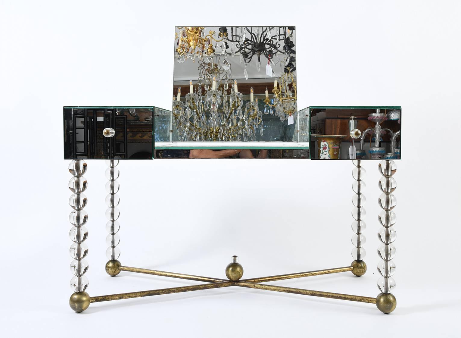 An Art Deco dressing table, mirrored on all four sides, the legs made of solid glass balls joined with a brass entretoise topped with a glass finial, the middle section opening as a dressing mirror and flanked by two drawers with glass