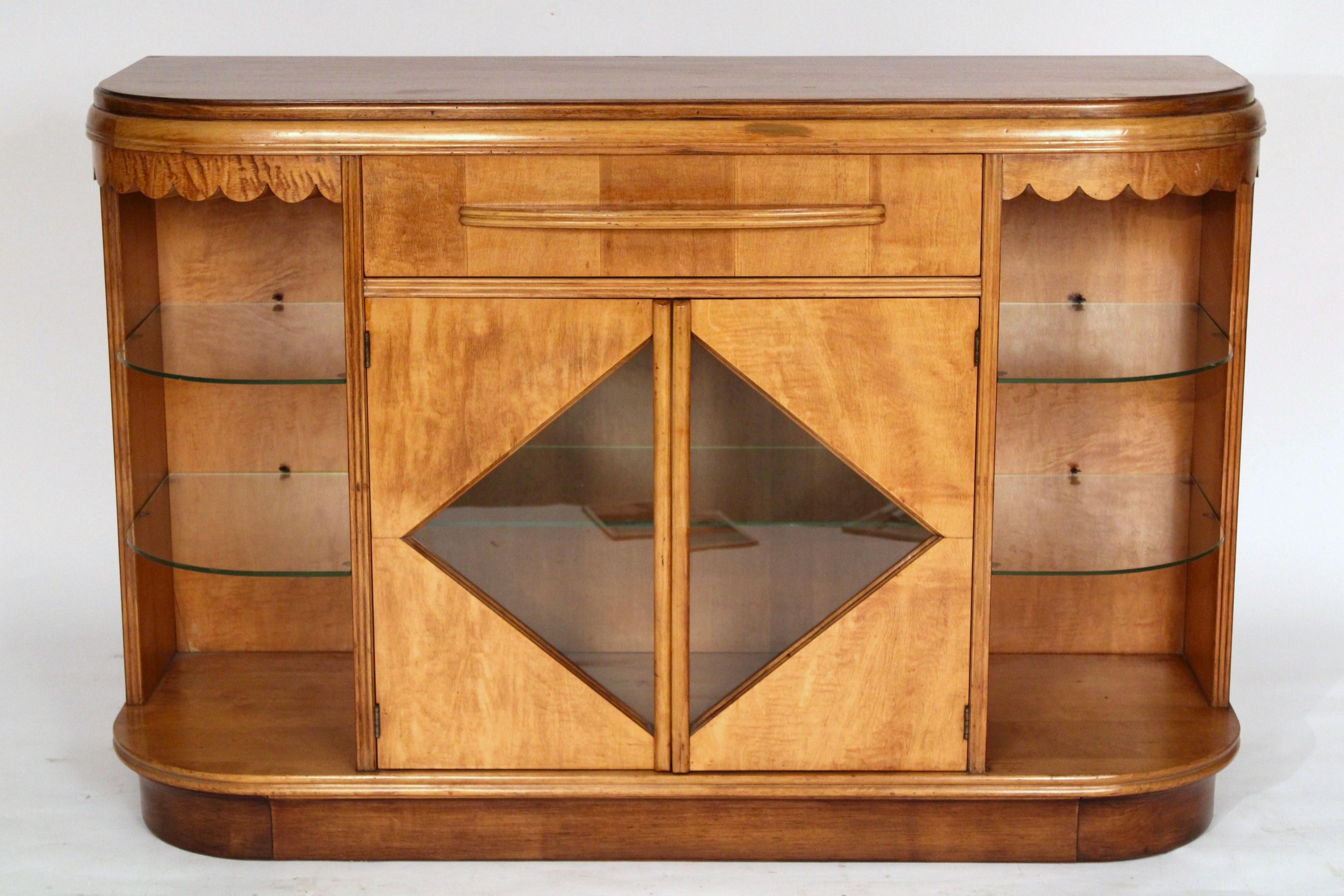 Art Deco mixed wood side cabinet, circa 1950's. With an elongated demi lune shaped  top, a central drawer over two doors with diamond shaped glass doors, sides with 2 glass shelves on each side, resting on a recessed walnut base. The woods used on