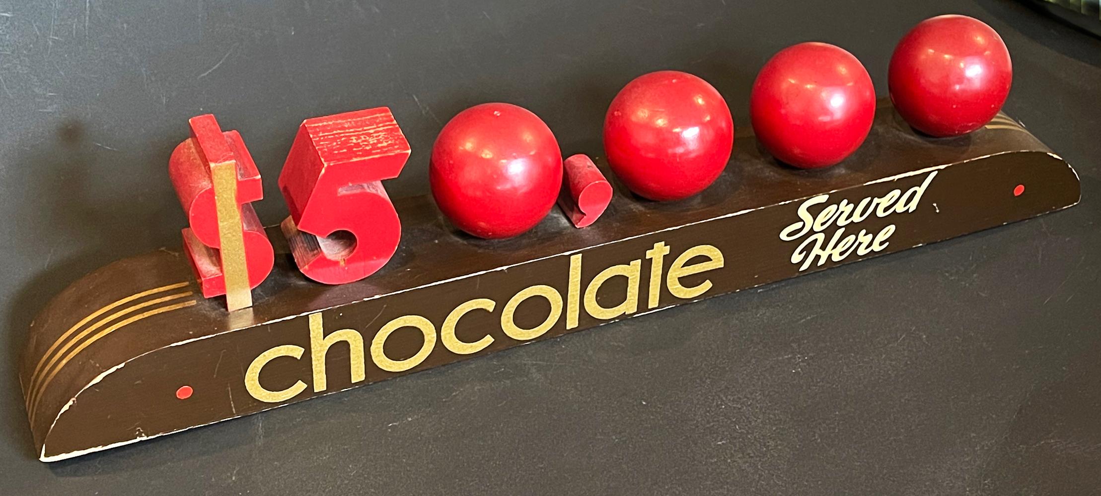 A remarkable example of sleek, streamlined Moderne design at the end of the Art Deco era, this painted wood sign for $50,000 Chocolate syrup is a very rare survivor. The designer used spherical wood balls to represent each of the 