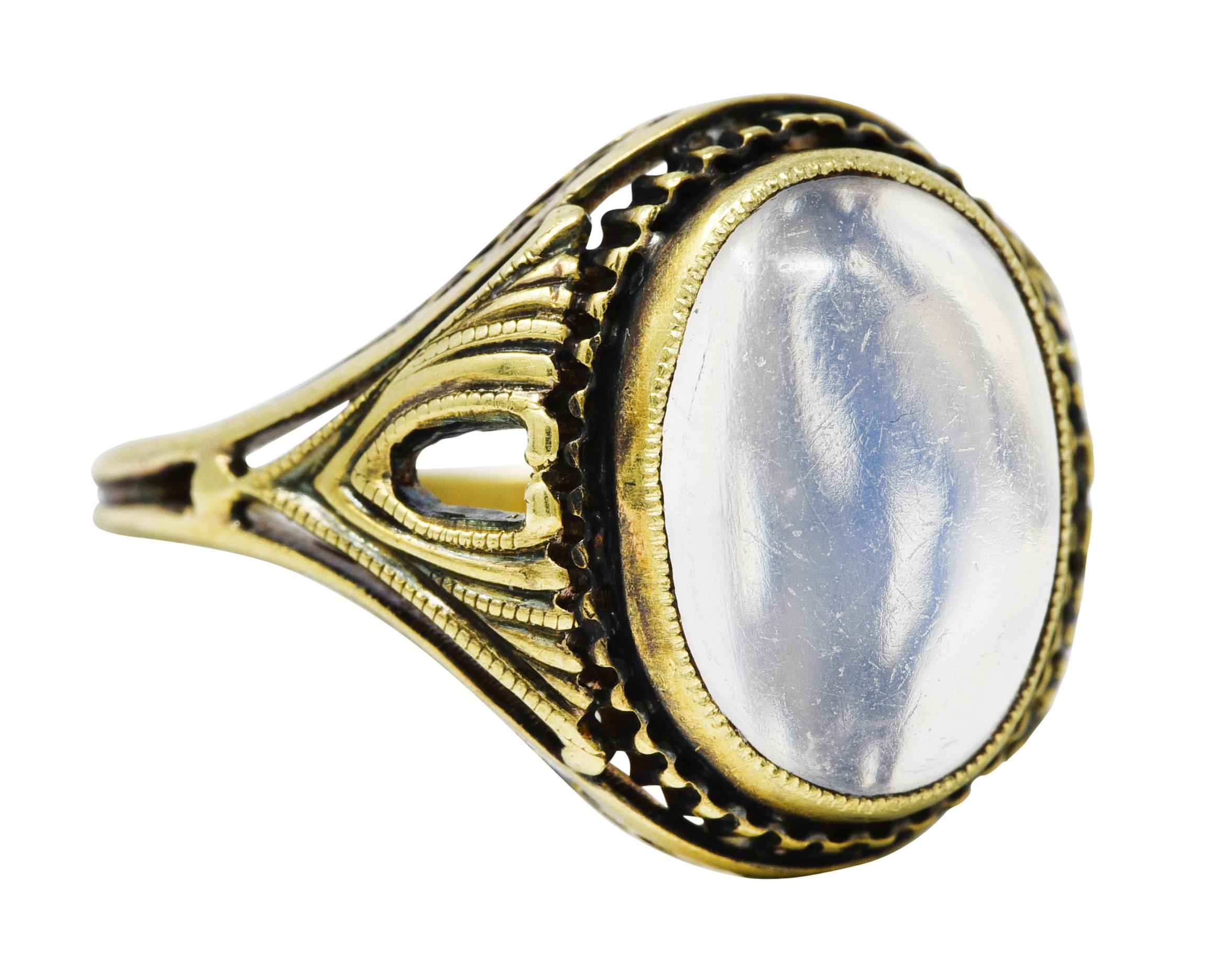 Ring centers a bezel set oval moonstone cabochon measuring 8.0 x 11.0 mm. Translucent and colorless with blue adularescence. With a grooved surround and sweeping ridged shoulders. Accented by lattice profile. Stamped 14 for 14 karat gold. With