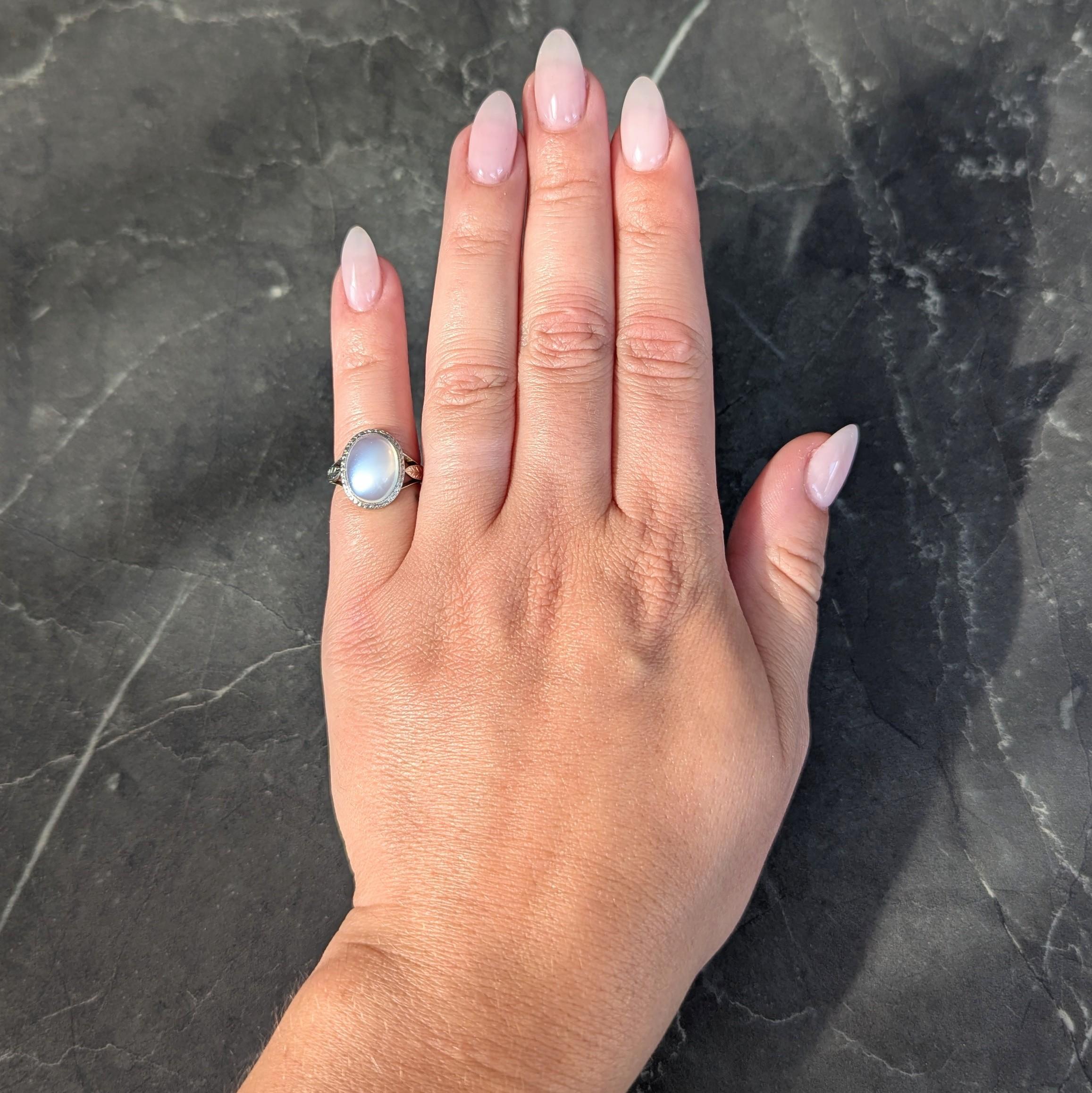 Centering an oval shaped moonstone cabochon measuring 12.0 x 9.0 mm
Translucent colorless body and displaying white adularescence
Flush set with engraved scroll motif halo and pierced profile
Flanked by tri-split shoulders engraved with wheat