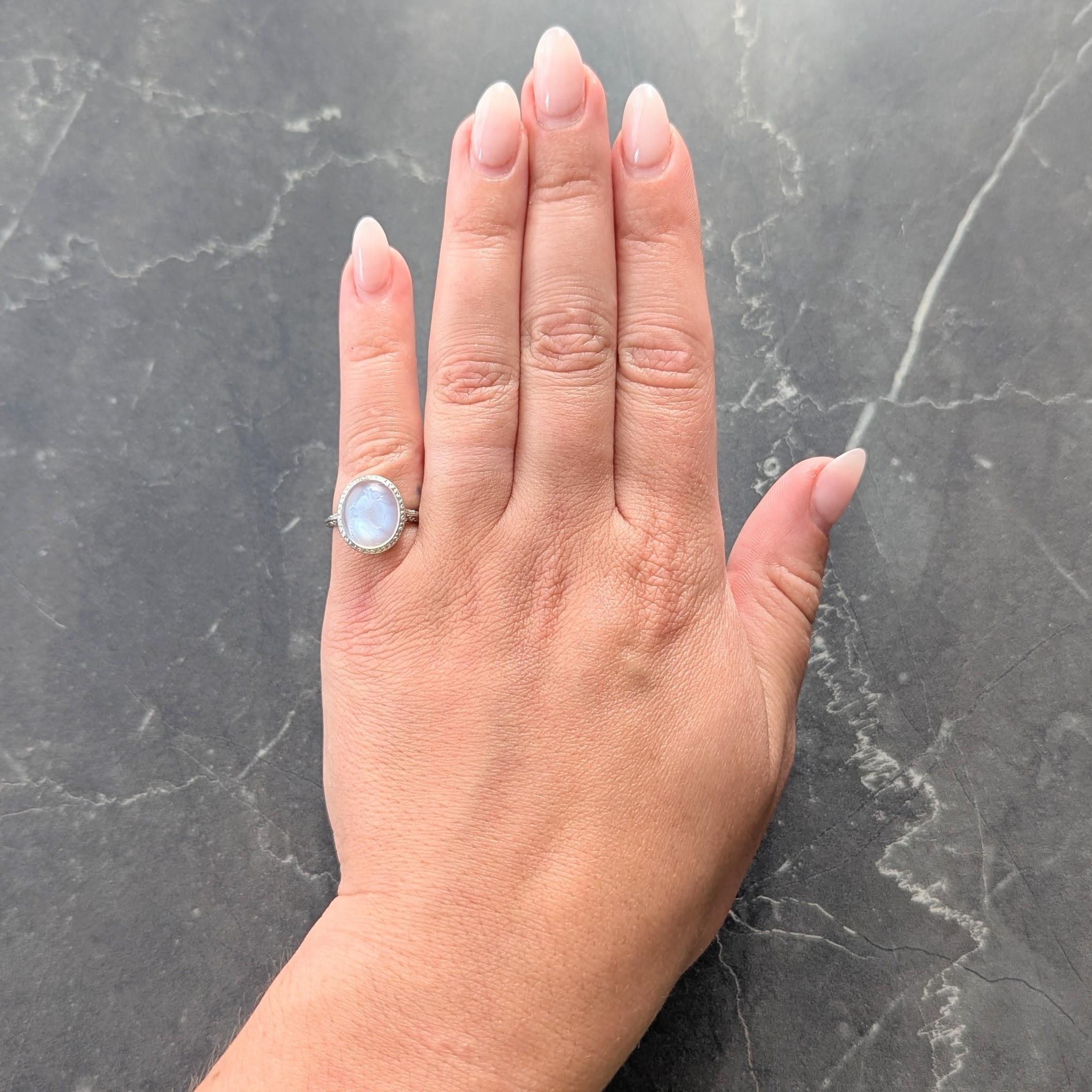 Centering an oval shaped moonstone cabochon measuring 11.5 x 9.5 mm
Translucent colorless body with vivid blue adularescence - flush set
Carved with an intaglio of a woman in profile with headpiece
Featuring an engraved halo surround
With a pierced