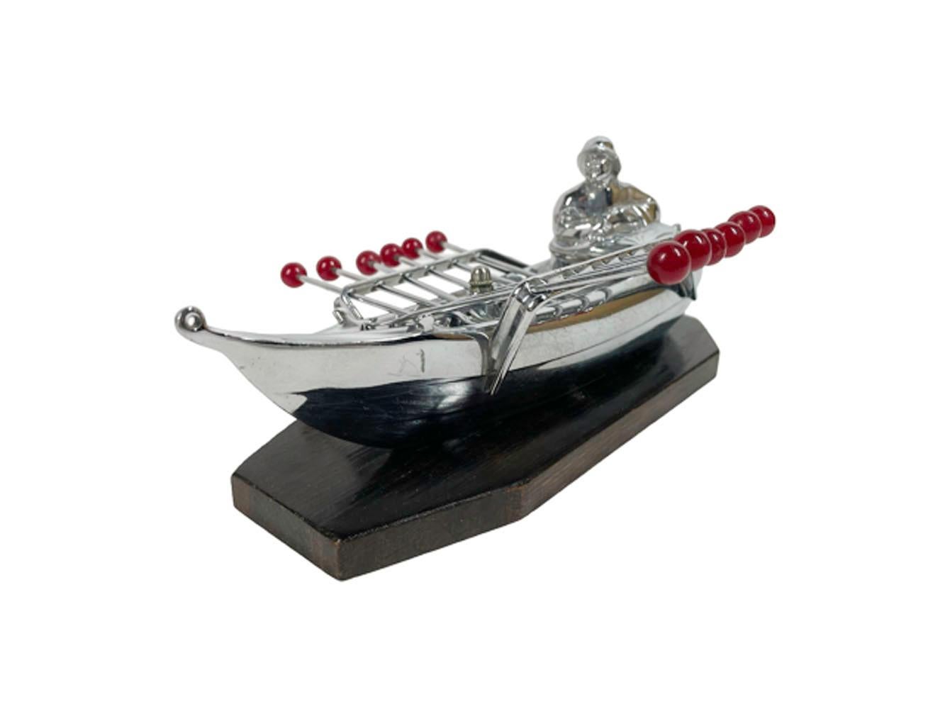 Art Deco chromed cast metal cocktail pick holder in the form of a man wearing foul weather gear in a small boat with perforated rails holding 12 forked picks with red ball ends and all set on a wood base.