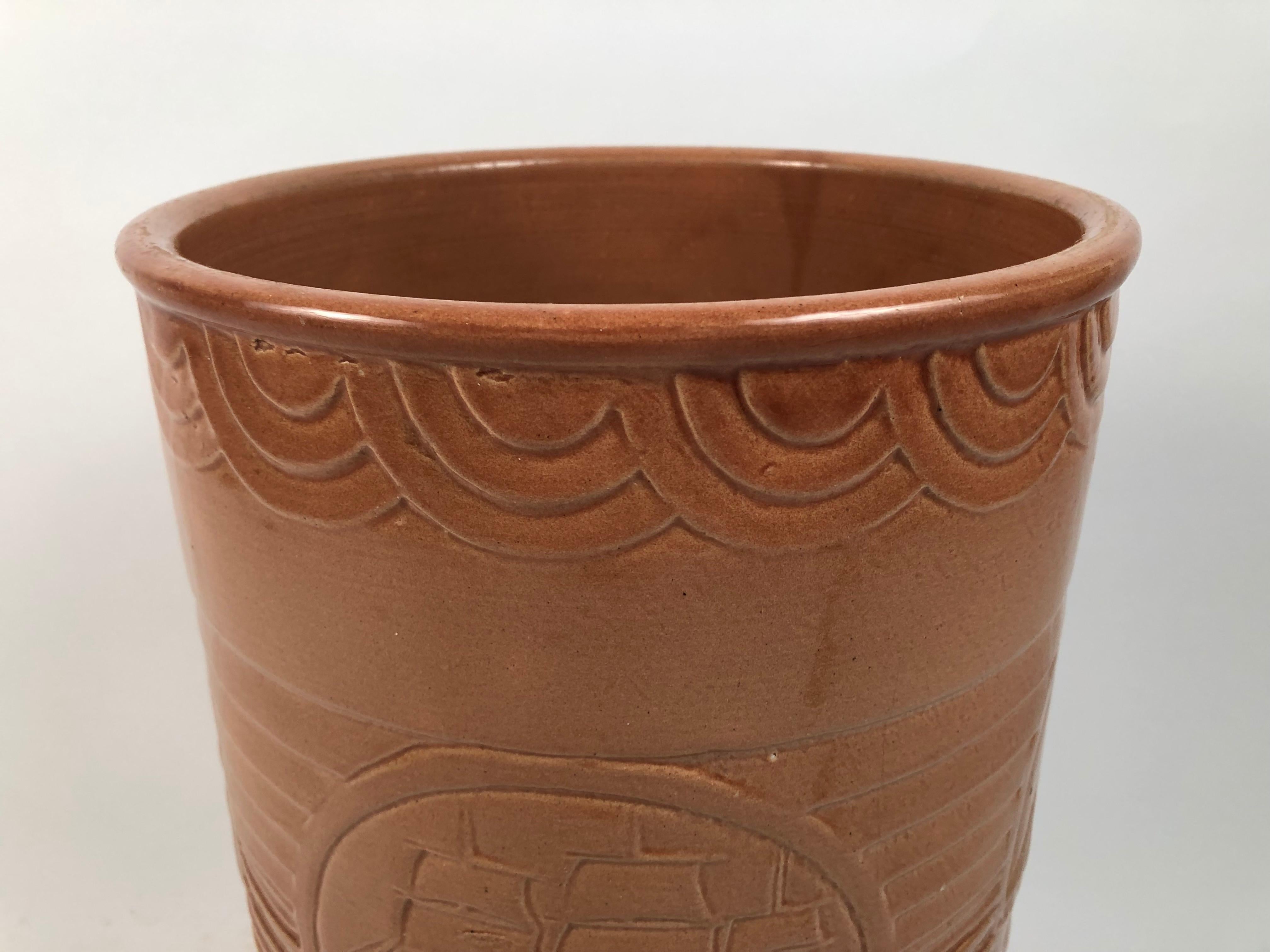 An American Art Deco period glazed tan ceramic waste basket, of cylindrical form, decorated with a bas relief medallion featuring a schooner in the center flanked by horizontal bands, with semi-circular geometric borders at the top and bottom. Would