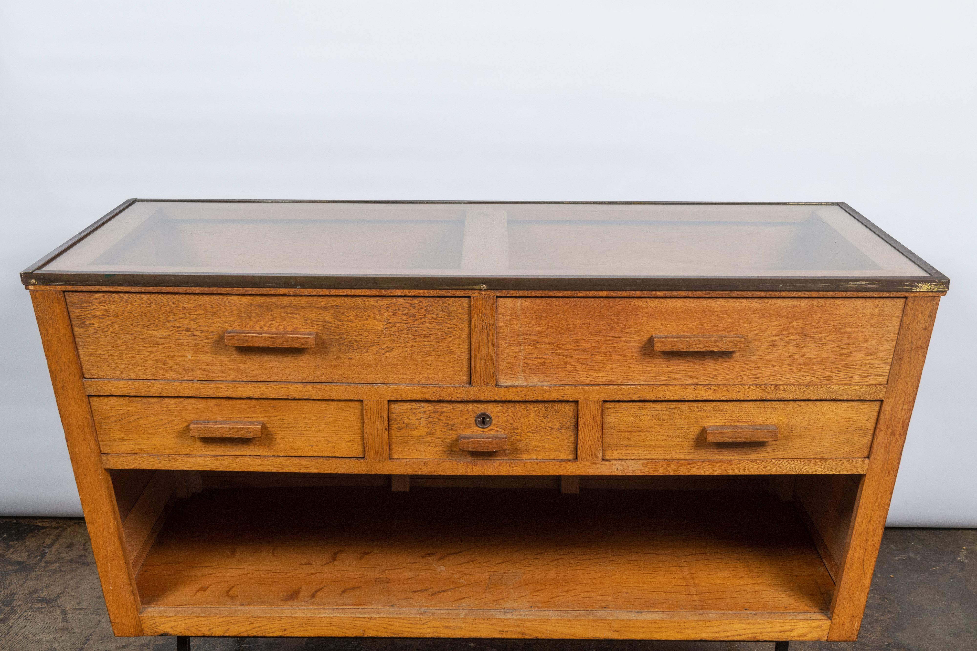 This vintage Art Deco cabinet in solid oak has a trapezoid shape, with two large drawers beneath a glass top trimmed in brass, three smaller drawers beneath, and a large storage space below, which sits upon metal legs. Great for displaying jewelry,