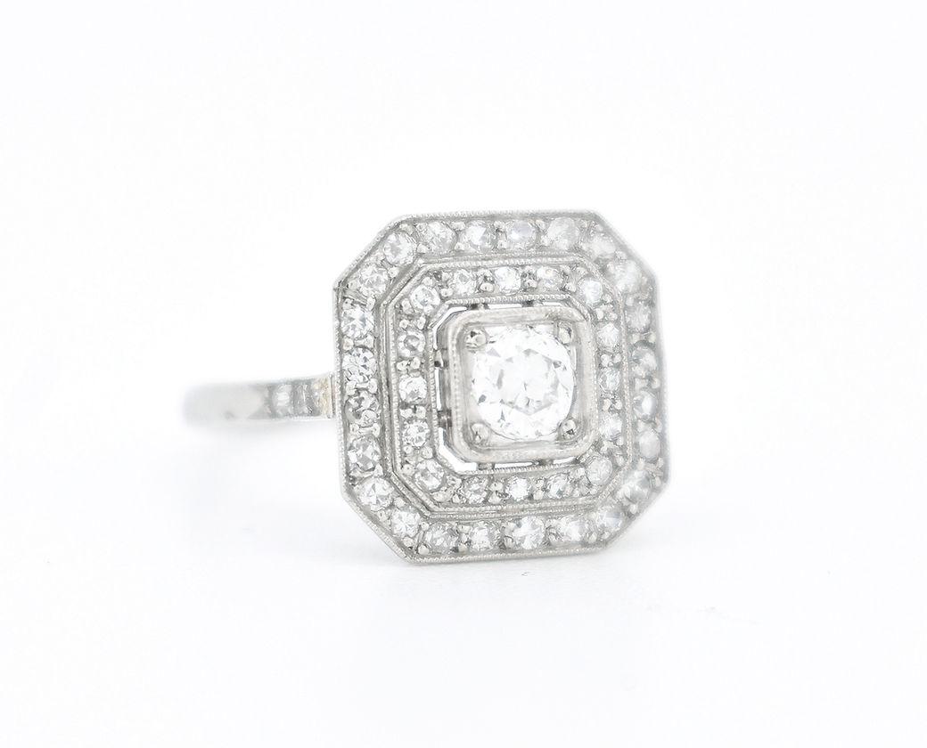 This exquisite Art Deco engagement ring, dating back to the 1920s, is crafted in platinum and showcases the timeless elegance of the era. At its center, it features an old-cut diamond of approximately 0.10 carats, surrounded by a striking double