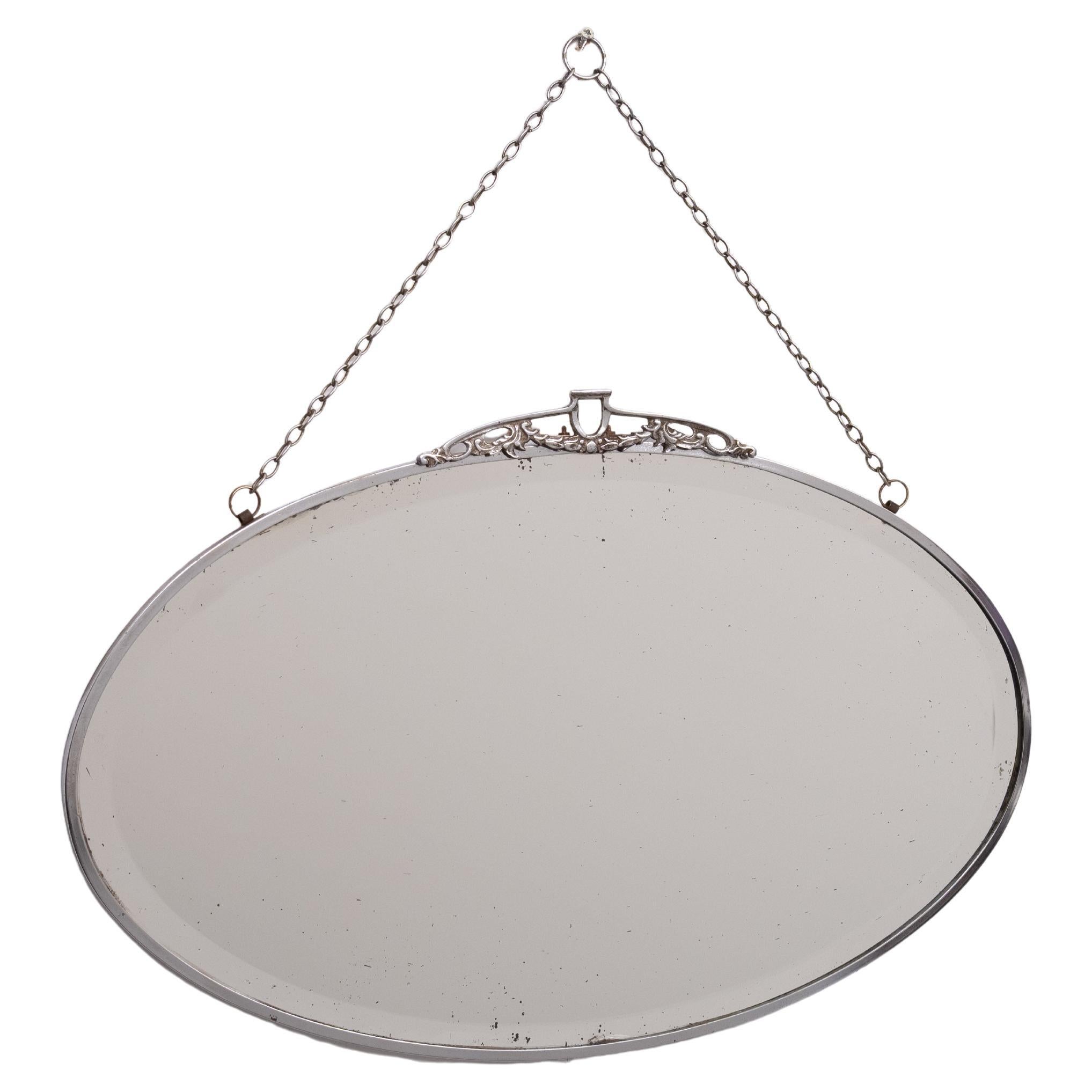 Miroir mural Art Deco de forme ovale. Livré avec son emballage d'origine. 
Chaîne . Miroir biseauté, cadre en nickel et ornement sur le dessus.                     est livrée avec juste ce qu'il faut de patine.
