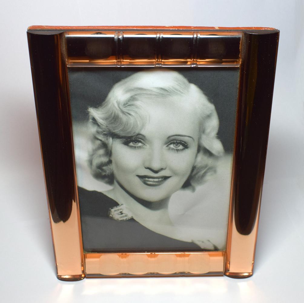 This 1930s Art Deco picture frame is an absolute delight and is In exceptional condition. Features a very thick peach/rose colored mirror glass with bevelled edges and design. Freestanding and can accommodate slightly larger than postcard size