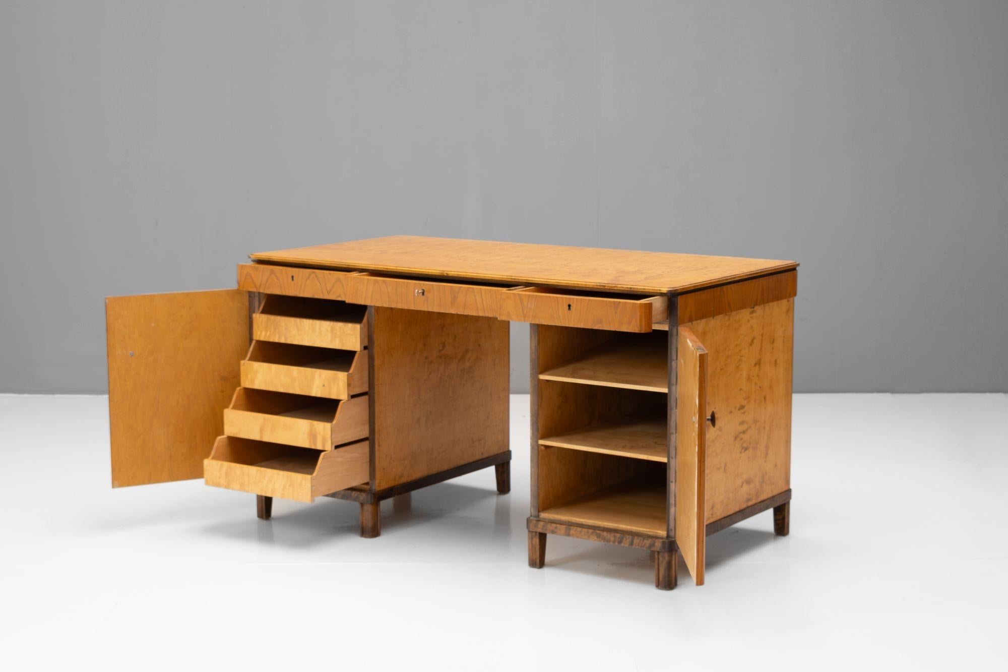Art-Deco-Schreibtisch des frühen 20. Jahrhunderts aus Ulme und Birke. Drei abschließbare Schubladen im Oberteil. Ein Sockel mit Schubladen und der andere mit drei festen Einlegeböden. Schweden um 1930.

H 76cm (KH 66cm) x B 136cm (KH 49cm) x T 70cm