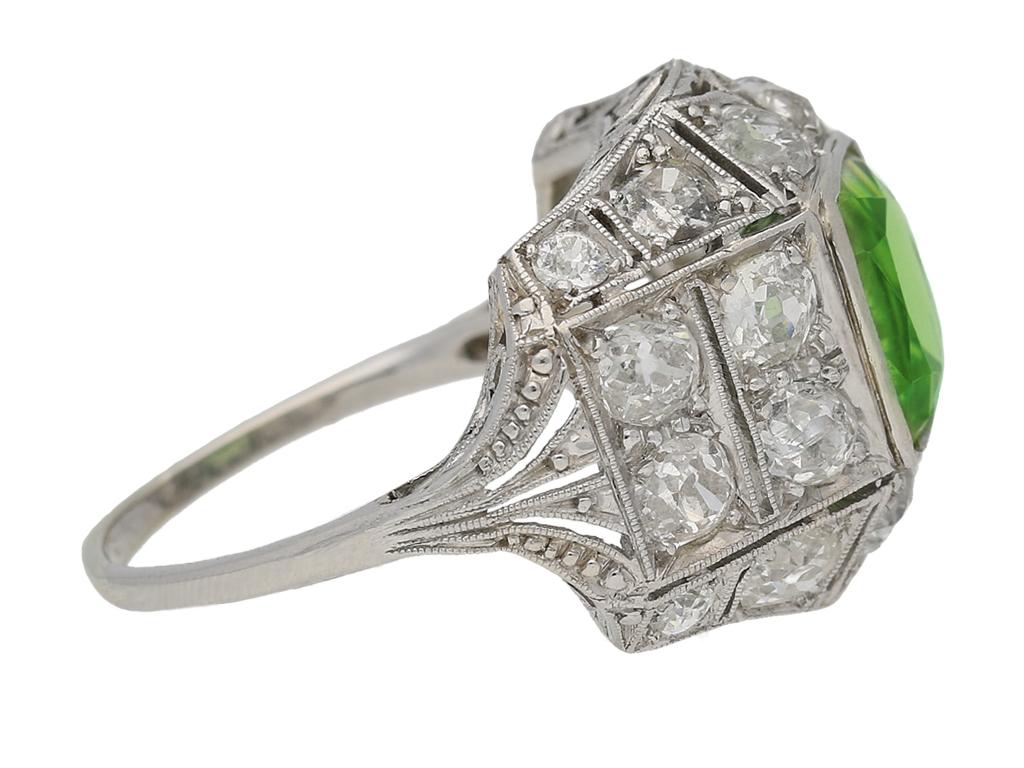Art Deco peridot and diamond cluster ring. Set with a cushion shape old cut natural peridot to centre in an open back rubover setting with an approximate weight of 4.50 carats, further set with twenty cushion shape old cut diamonds in open back