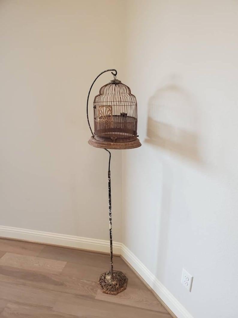 A rustically elegant antique bird cage, circa 1920s, architectural shaped dome, period Art Deco elements, hanging from tall wrought iron shepard hook curved stand with charming cast iron figural bird motif. Perfectly patinated, remnants of the