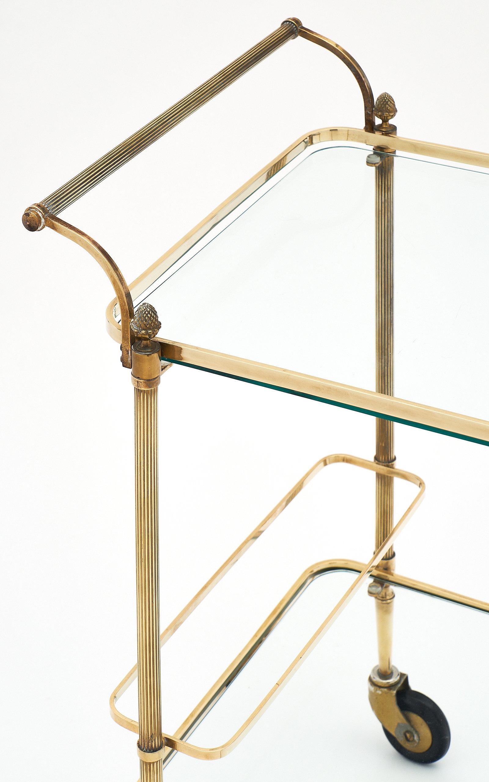 French Art Deco period bar cart with finials. This elegant, simple, and very sturdy piece features two shelves, a handle, decorative finials, and a bottle holder. This is a charming French bar cart we couldn’t resist. It is made of brass with glass