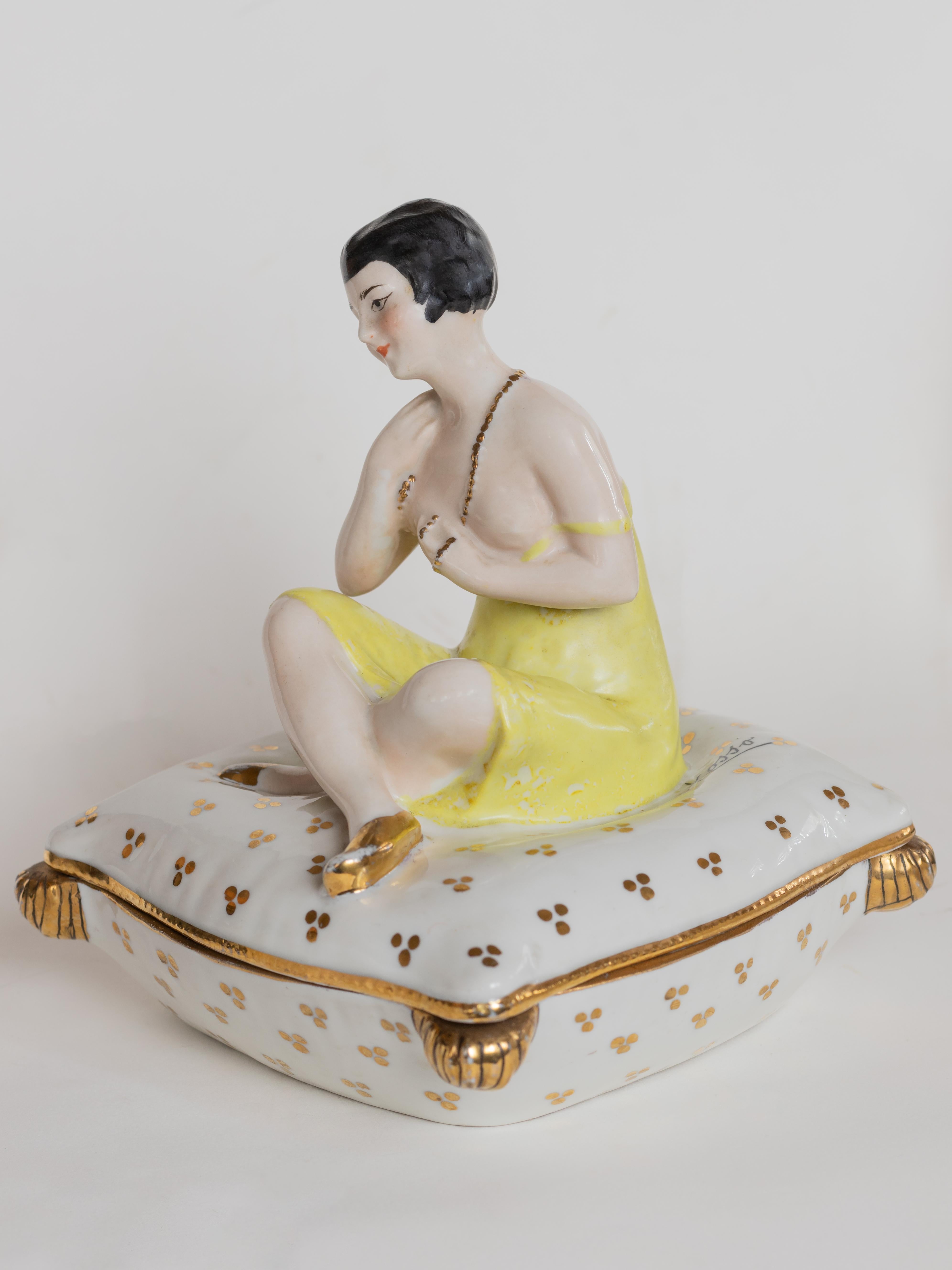 A beautifully crafted Art Deco powder box or jewelry box, adorned with a delicate figurine of a woman in a yellow dress, resting on a plush cushion sofa. The piece bears the mark 