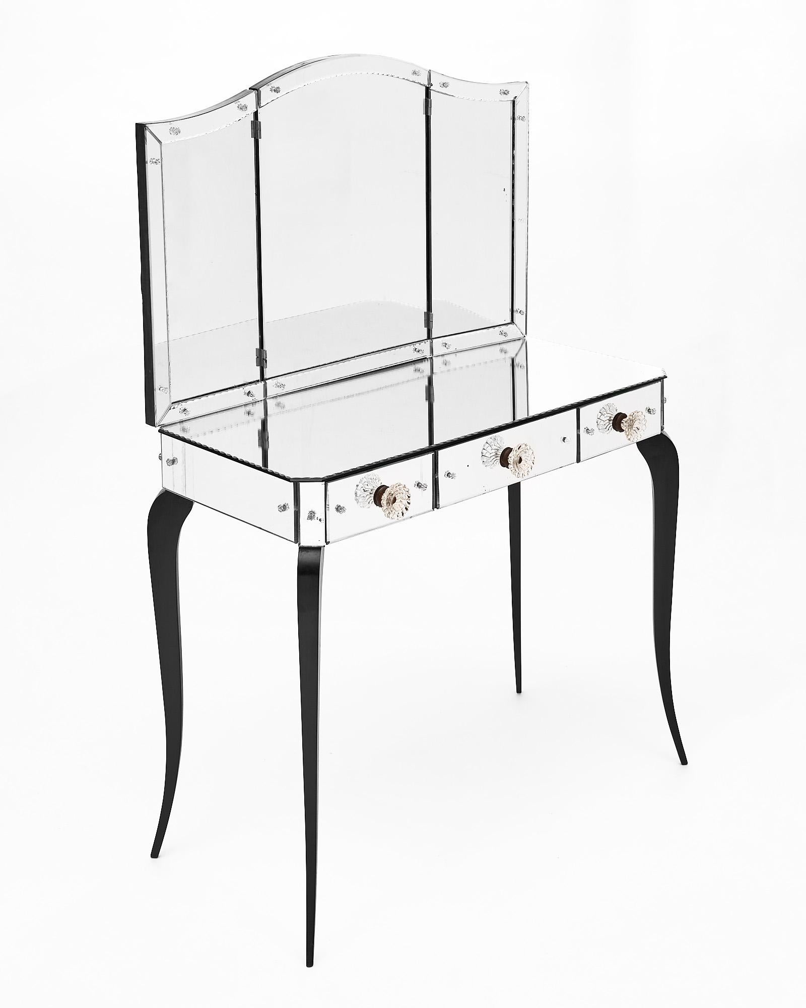 Vanity from France made with a beveled glass veneer and three drawers with original glass pulls. The top has a hinged three part mirror. Fully open, the mirror has a width of 34.25”. The height to the top of the vanity table is 30.5”. From the floor