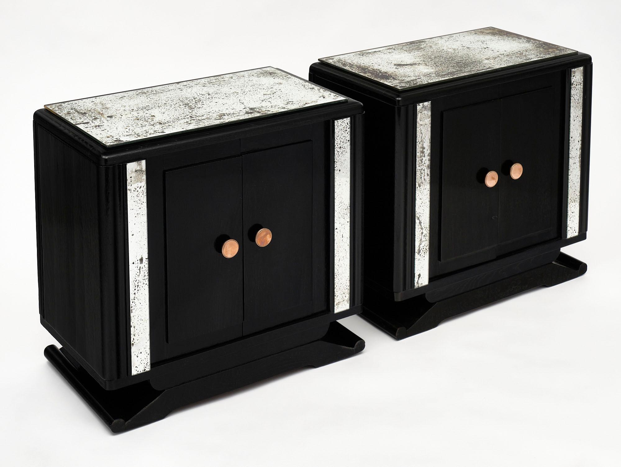 A pair of Art Deco period vintage side tables from France with antique mirrored tops and side accent pieces. There are two doors on each featuring brass knobs that open to a compartment with one interior shelf. We love the strong Deco lines of this