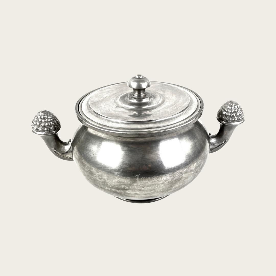 20th Century, Sweden. This Art Deco pewter terrine with lid by GAB features arms decorated with grape-shaped handles. The front is adorned with a relief of a man and woman, while the backside is engraved with a Swedish gift message. The piece is in