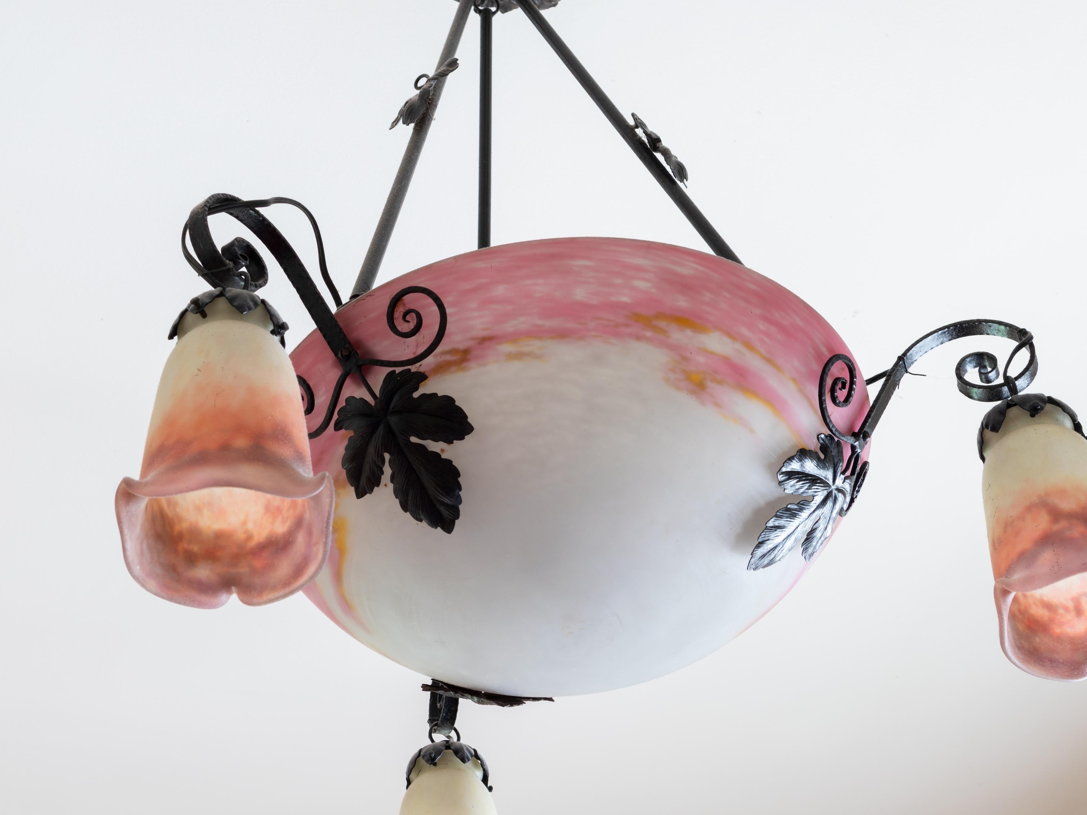 This Art Deco chandelier by Muller Freres features a central pink glass bowl shade with elegant wrought iron fitments supporting three satellite shades. 

The ironwork rods are beautifully decorated with grape leaves, adding a touch of