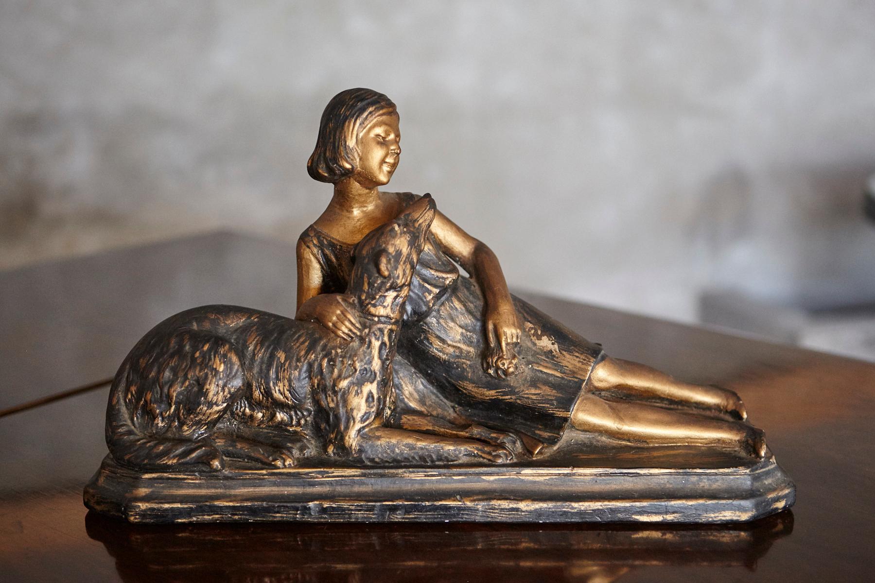 Scultura d'epoca in gesso raffigurante una donna seduta con il suo cane, probabilmente risalente al periodo Art Déco. Il pezzo presenta linee pulite e una composizione semplice ed elegante tipica dell'epoca. Rifinita nei toni del nero e dell'oro,